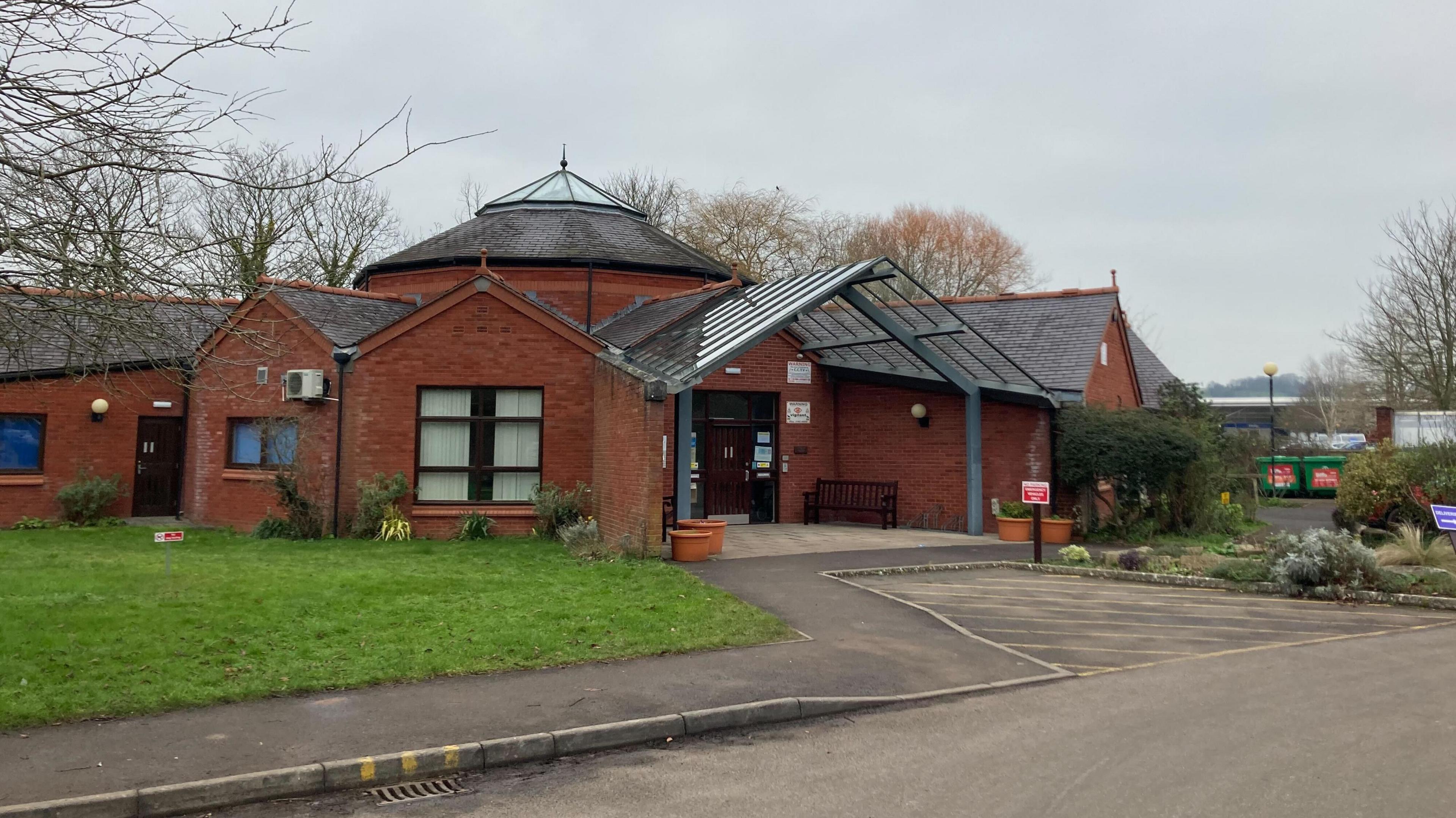 The picture shows the outside of Langport surgery.  There are trees in the background and the picture was taken on a cloudy day.