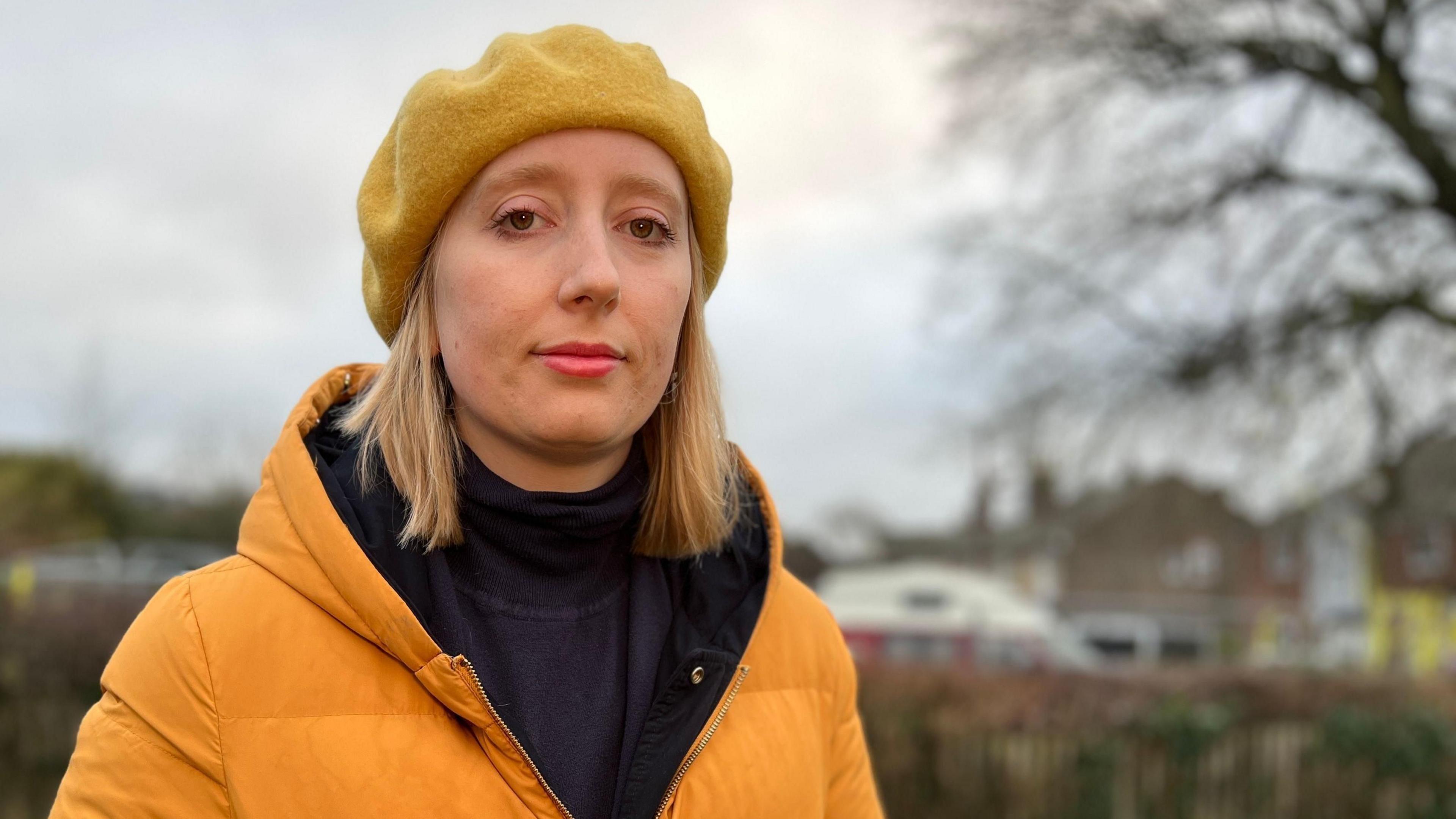 Kat Scott, a Wivenhoe resident, stood in a park with a tree behind her. She is wearing an orange coat and an orange beret and a black polo neck jumper. She has bright lipstick on and is looking at the camera.