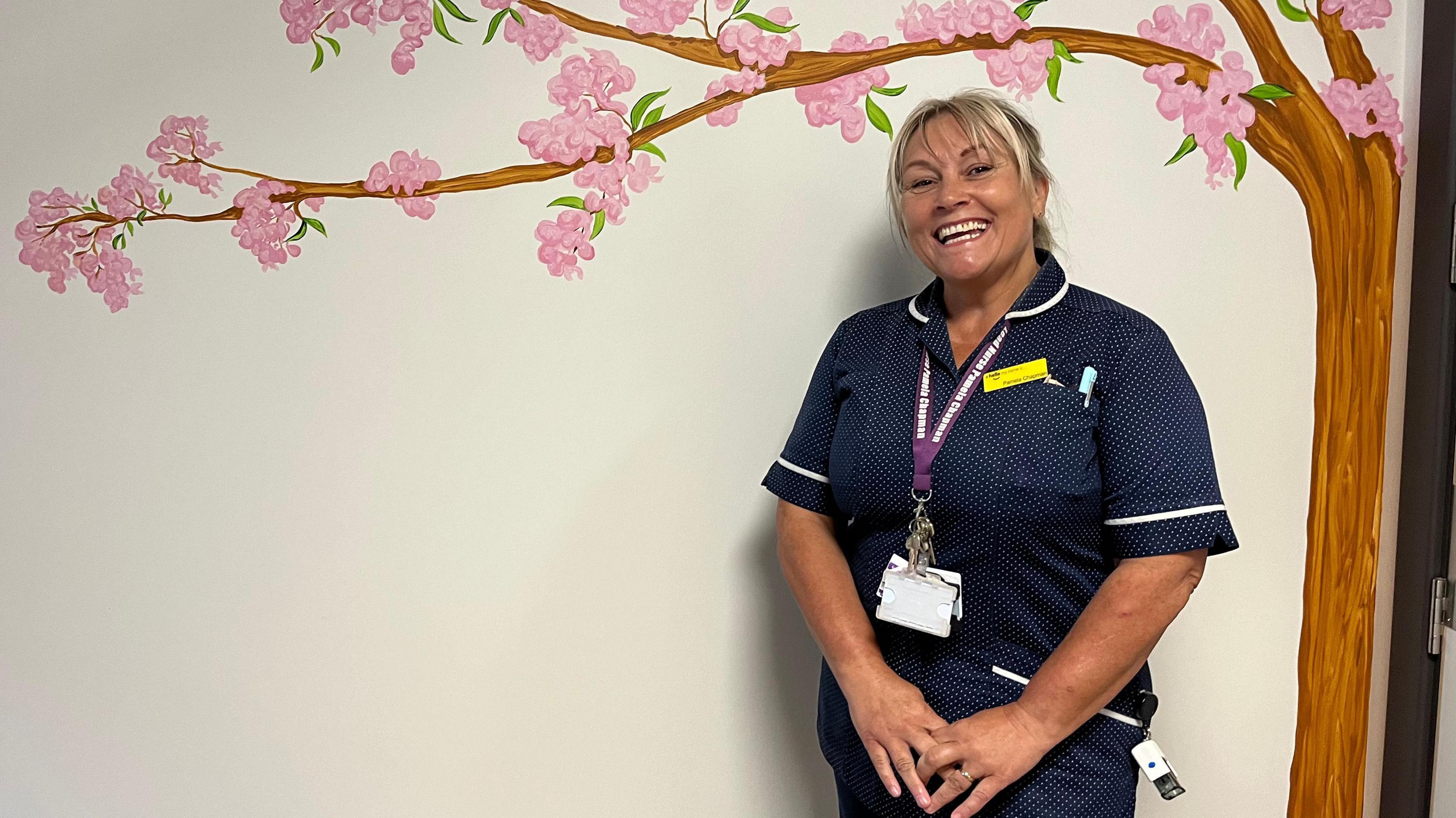 Lead Nurse for Palliative Care, Pamela Chapman, wearing navy scrubs and standing beside a white hospital wall which has a tree painted on it full of pink cherry blossom. She has blonde hair, a big smile and clasps her hands in front of her. 