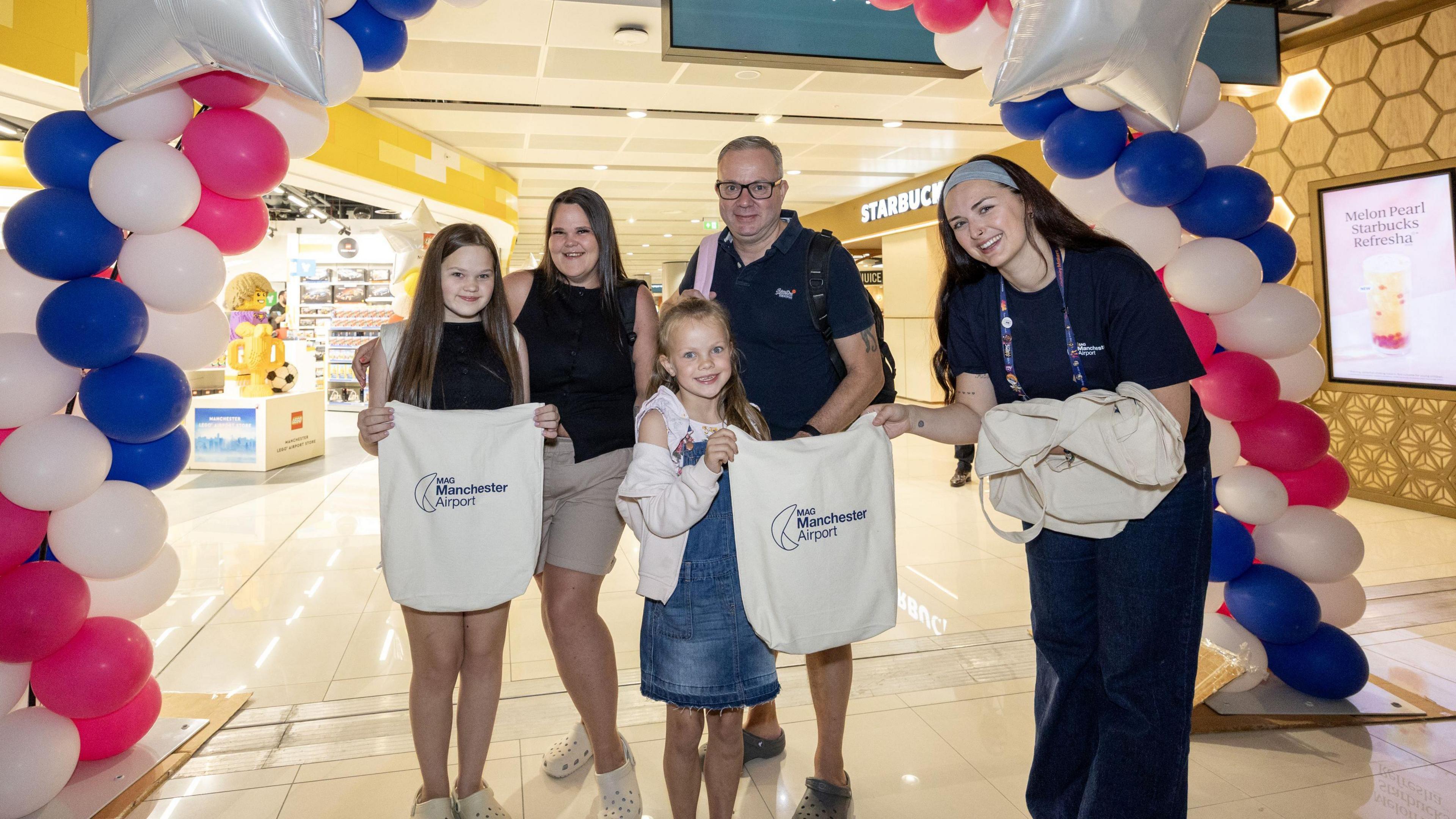 The Laxton family pose for a photo underneath a group of balloons celebrating the opening of the new Terminal 2 Section at Manchester Airport. The two young daughters are holding Manchester Airport-branded tote bags.