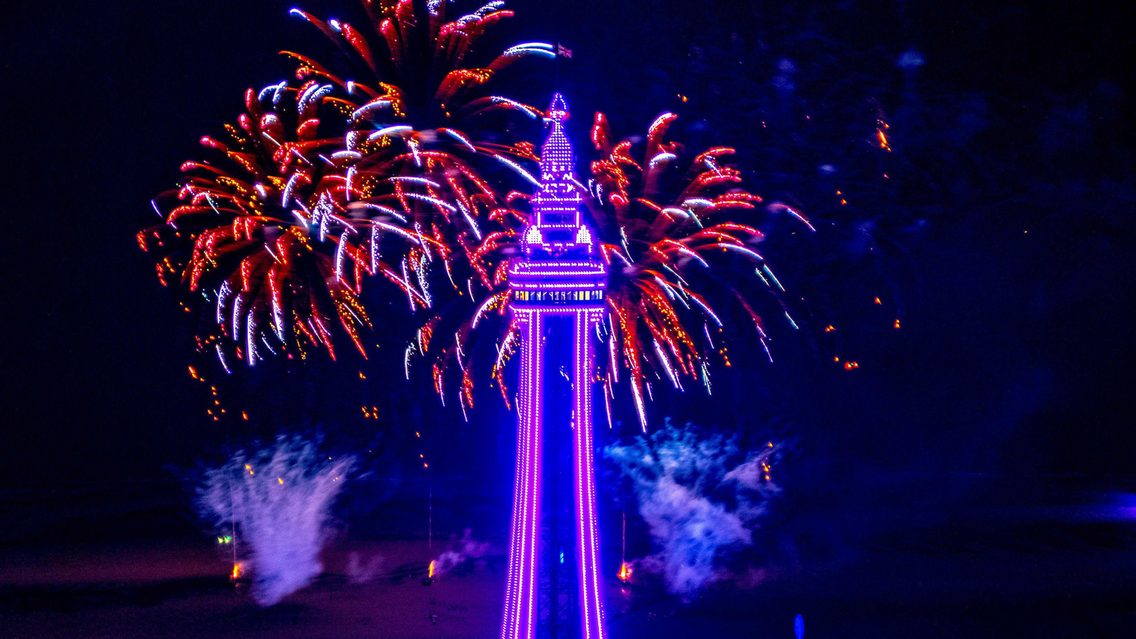 Blackpool Tower is lit up with light purple lights, with red fireworks exploding in the background.