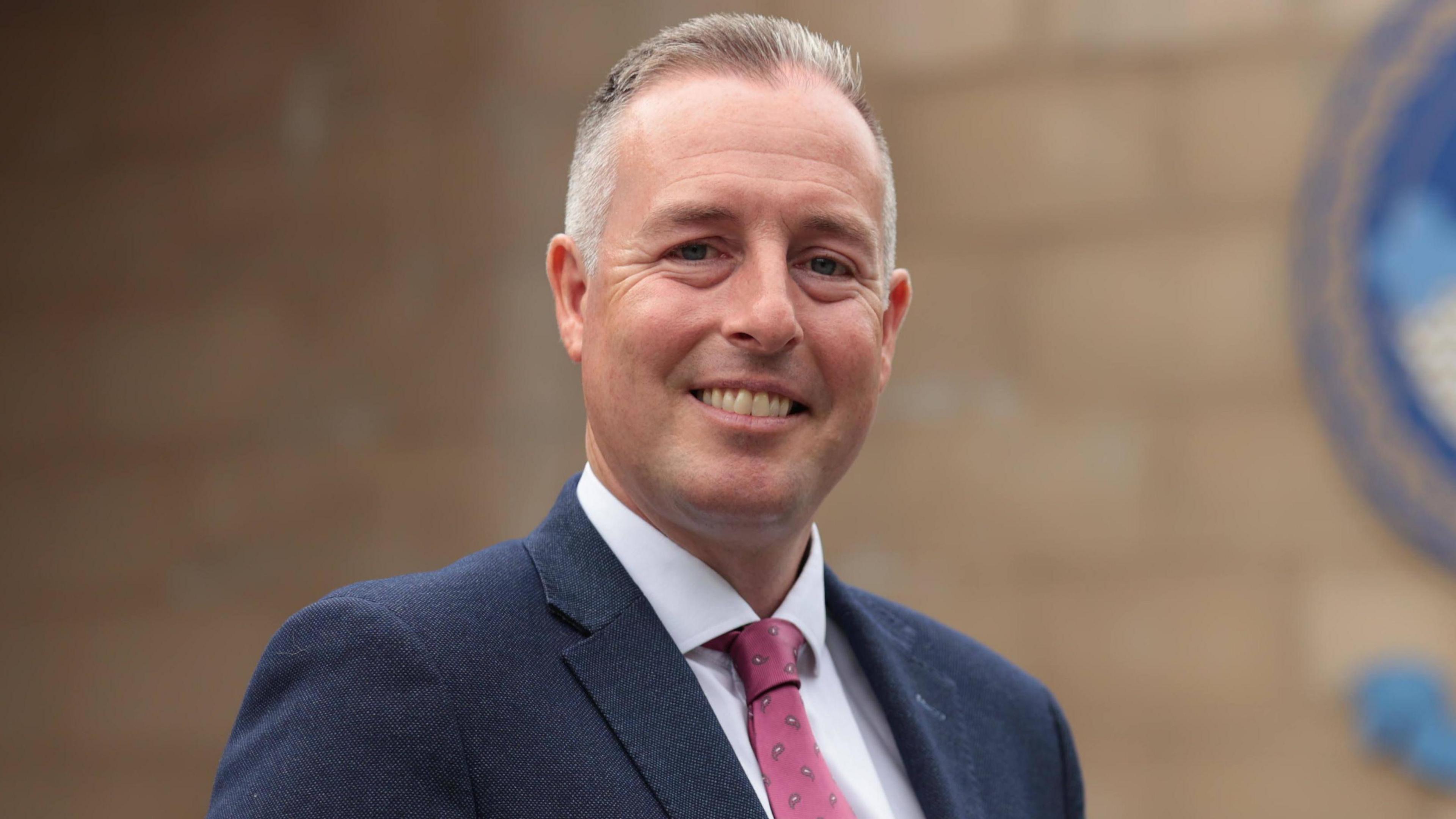 Paul Givan ;looking at the camera and smiling. He has a navy blazer and pink tie. 
