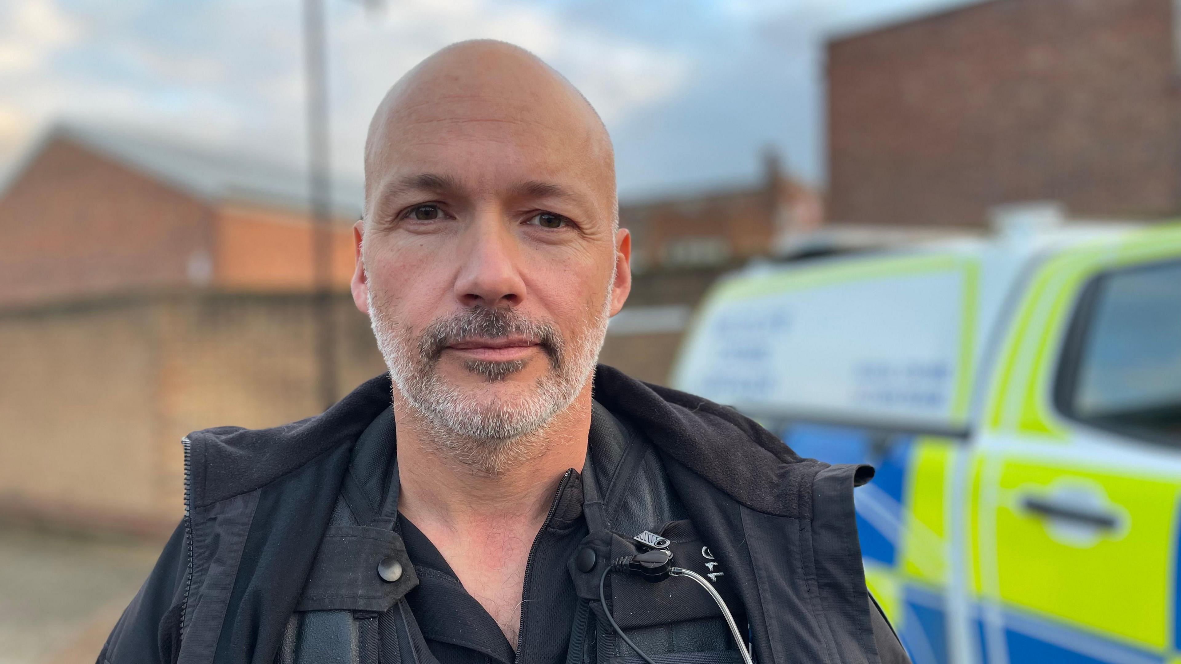 A head and shoulders shot of Det Con Aaron Flint. He is a bald man with a short grey beard. He is stood in front of a police van that is blurred in the background. He is wearing a black police uniform