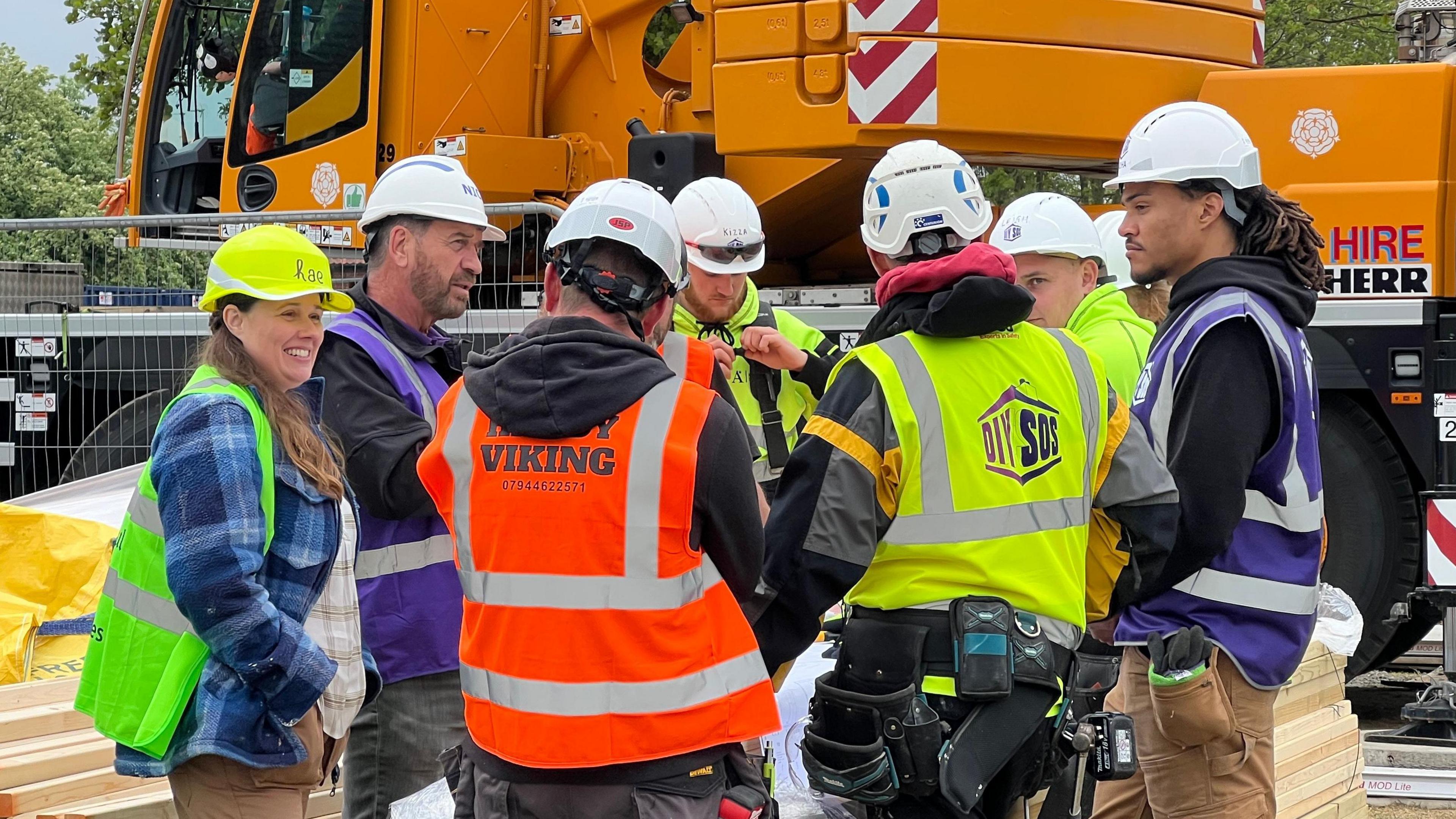 A group of seven tradespeople wearing hard hats and high-visibility clothing chat on a building site. A pile of timber is on the left of the image and a large yellow construction vehicle is visible in the background.