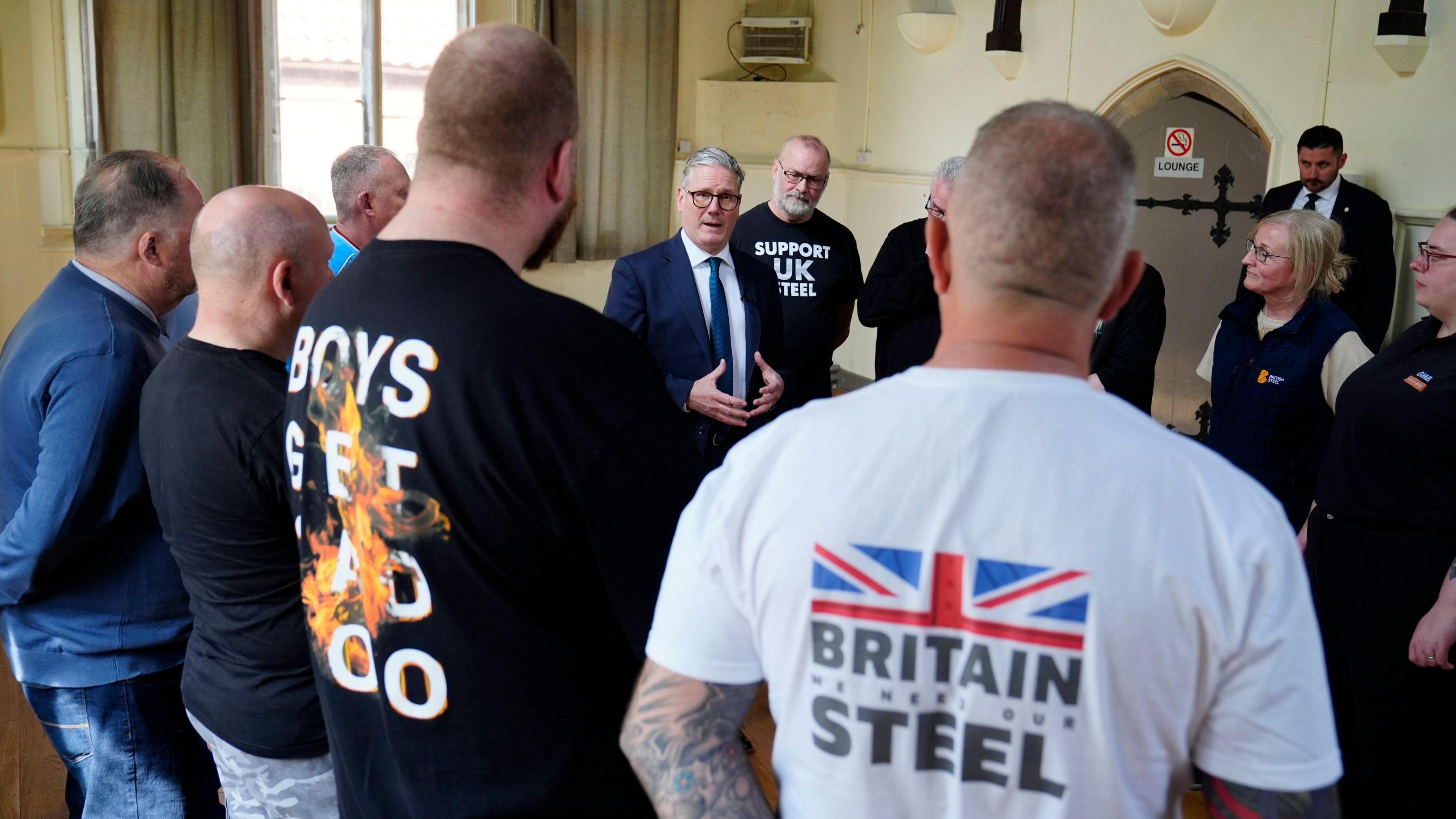 A group of people gathered in a village hall. In the middle of the group, a man in a suit and tie with glasses is addressing the group. Around him, there are eight men and two women- you can see a mixture of their faces and the backs of their head. The two men closest to the camera wear t-shirts reading 'BOYS GET SAD TOO' and 'BRITAIN WE NEED OUR STEEL'. The man closest to the speaker wears a 'SUPPORT UK STEEL' top.