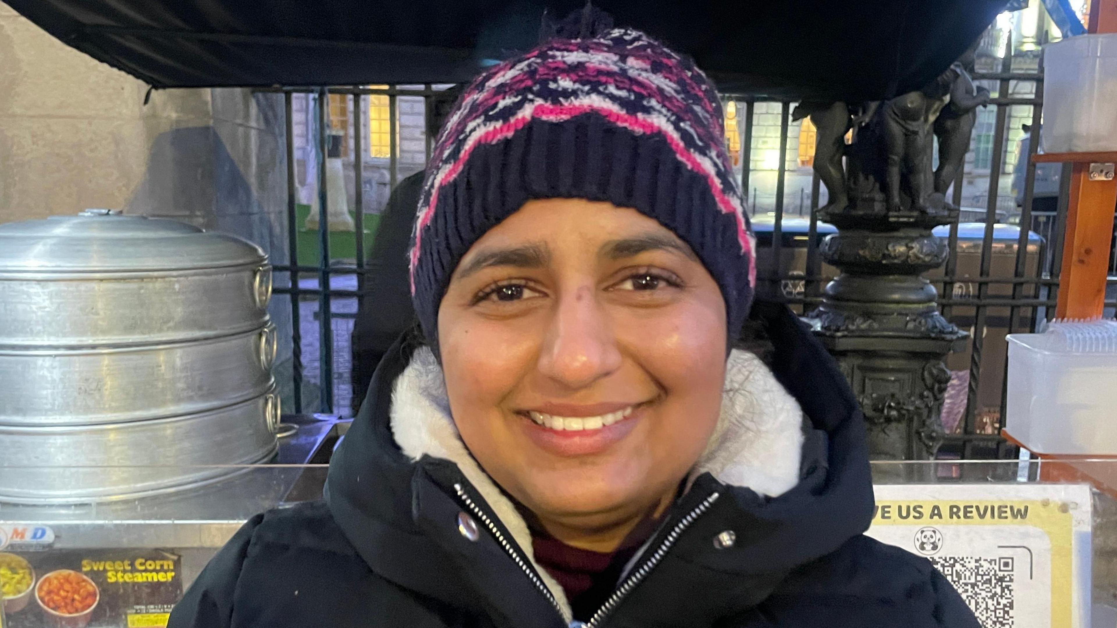 Anna is wearing a pink and purple hat and a black coat. She is standing outside Belfast City Hall. 