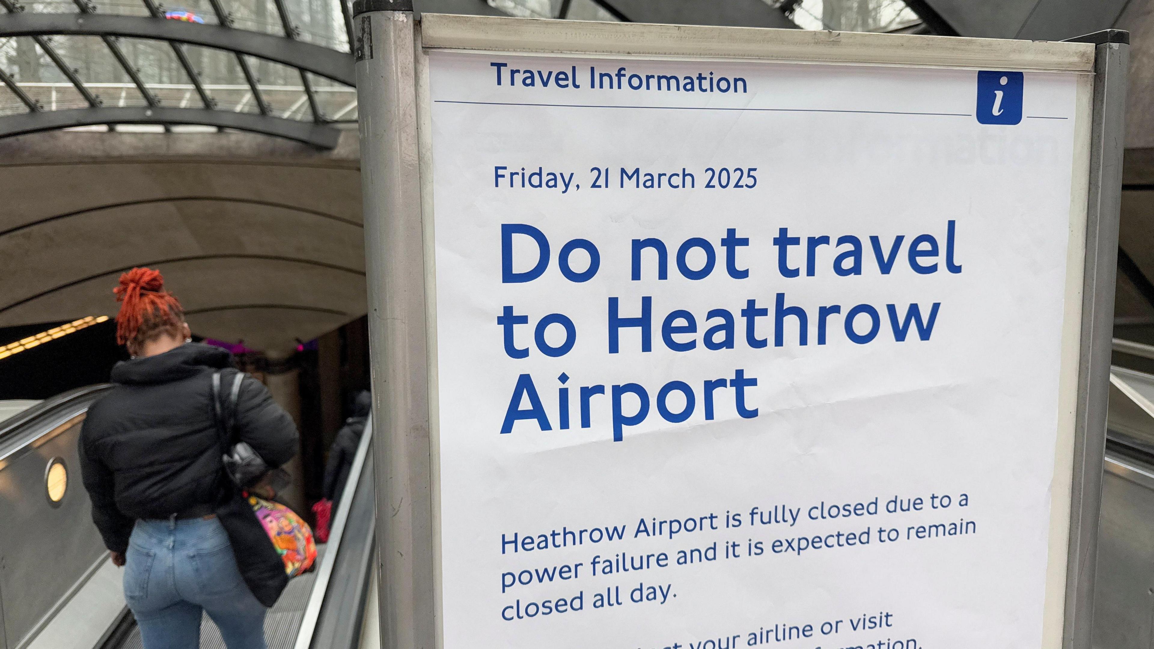 A travel information board with "Do not travel to Heathrow Airport" written in blue letters on it beside an escalator which has a woman in a black coat and blue jeans going down it