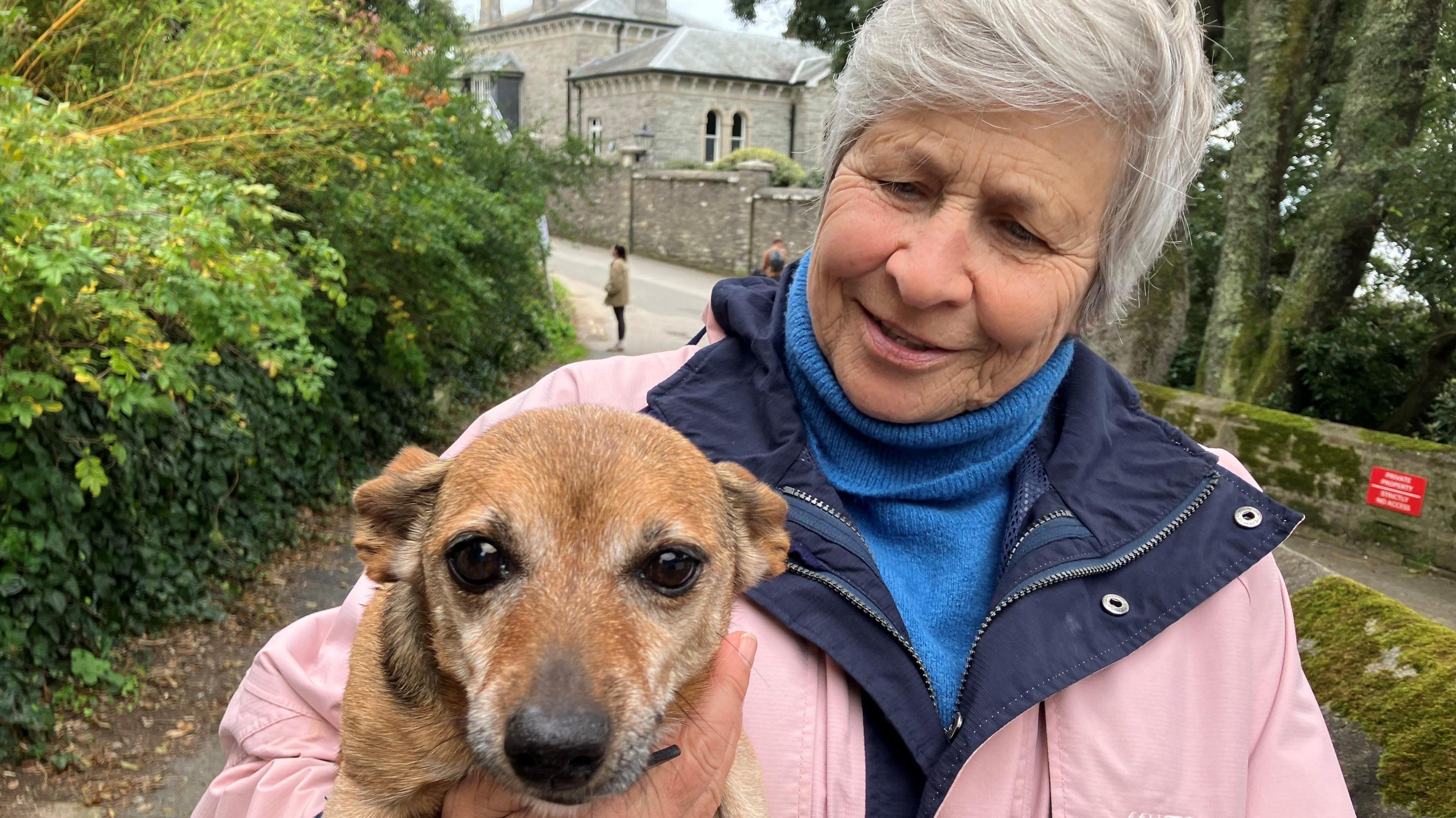 Woman with grey hair looking lovingly at her dog who she has in her arms. The dog is reaching out to the camera.