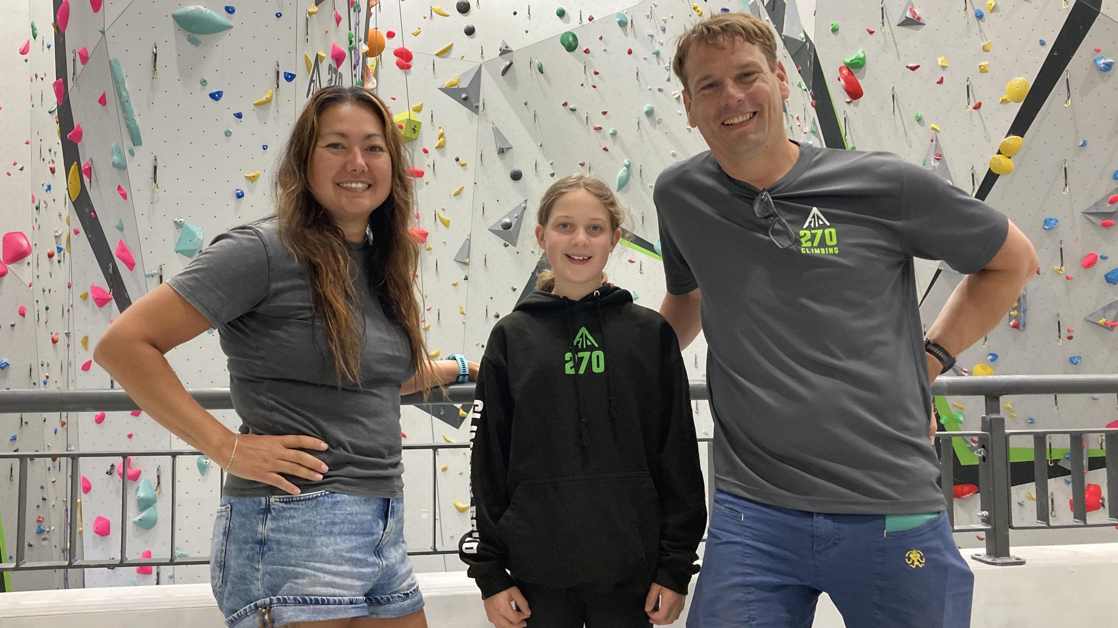 A woman and man stand with a young girl between them in front of a temporary climbing wall. The wall has dozens of coloured markers on it where people can put their hands and feet