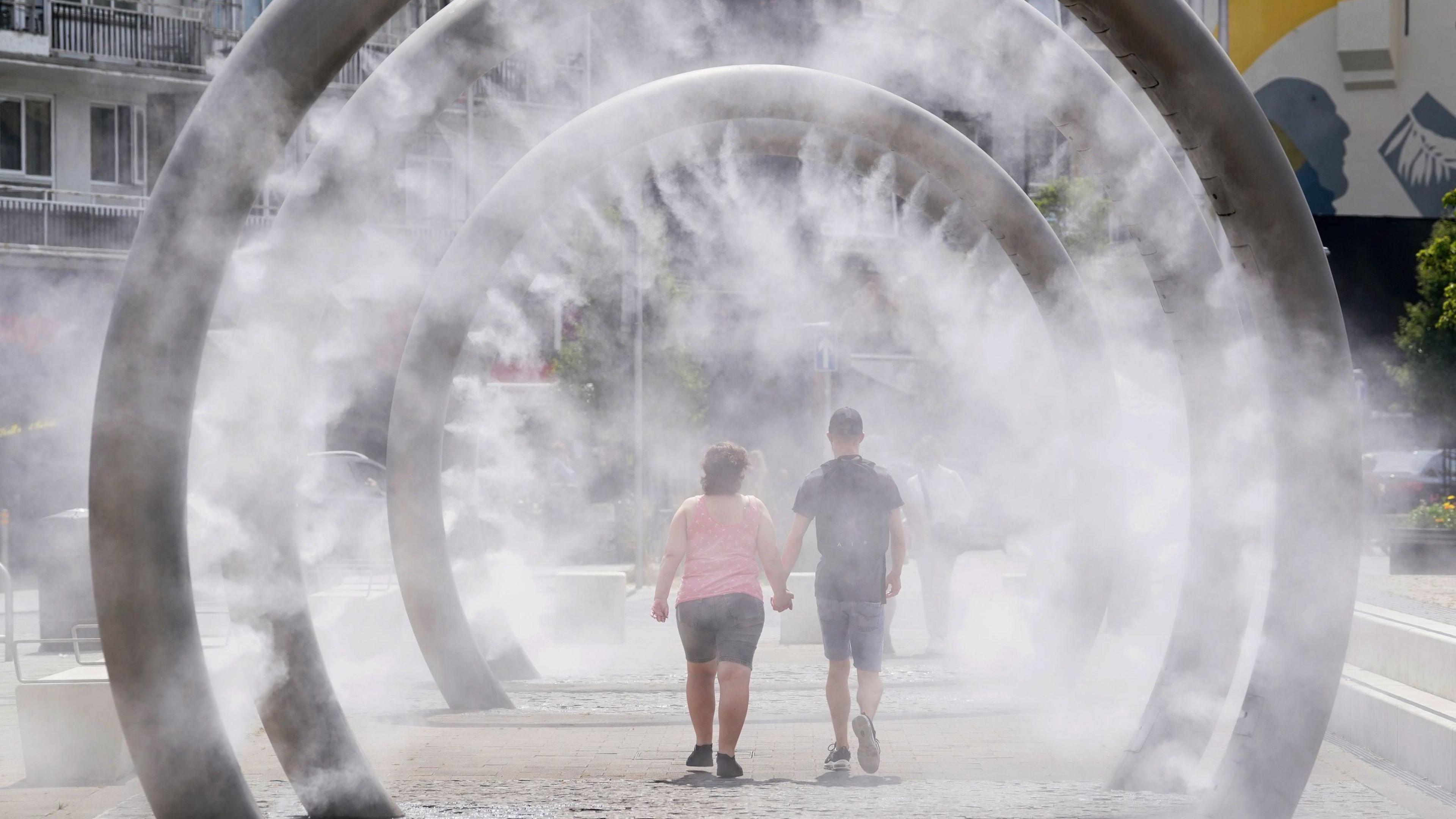 Two people holding hands walk through a water feature spraying a fine mist