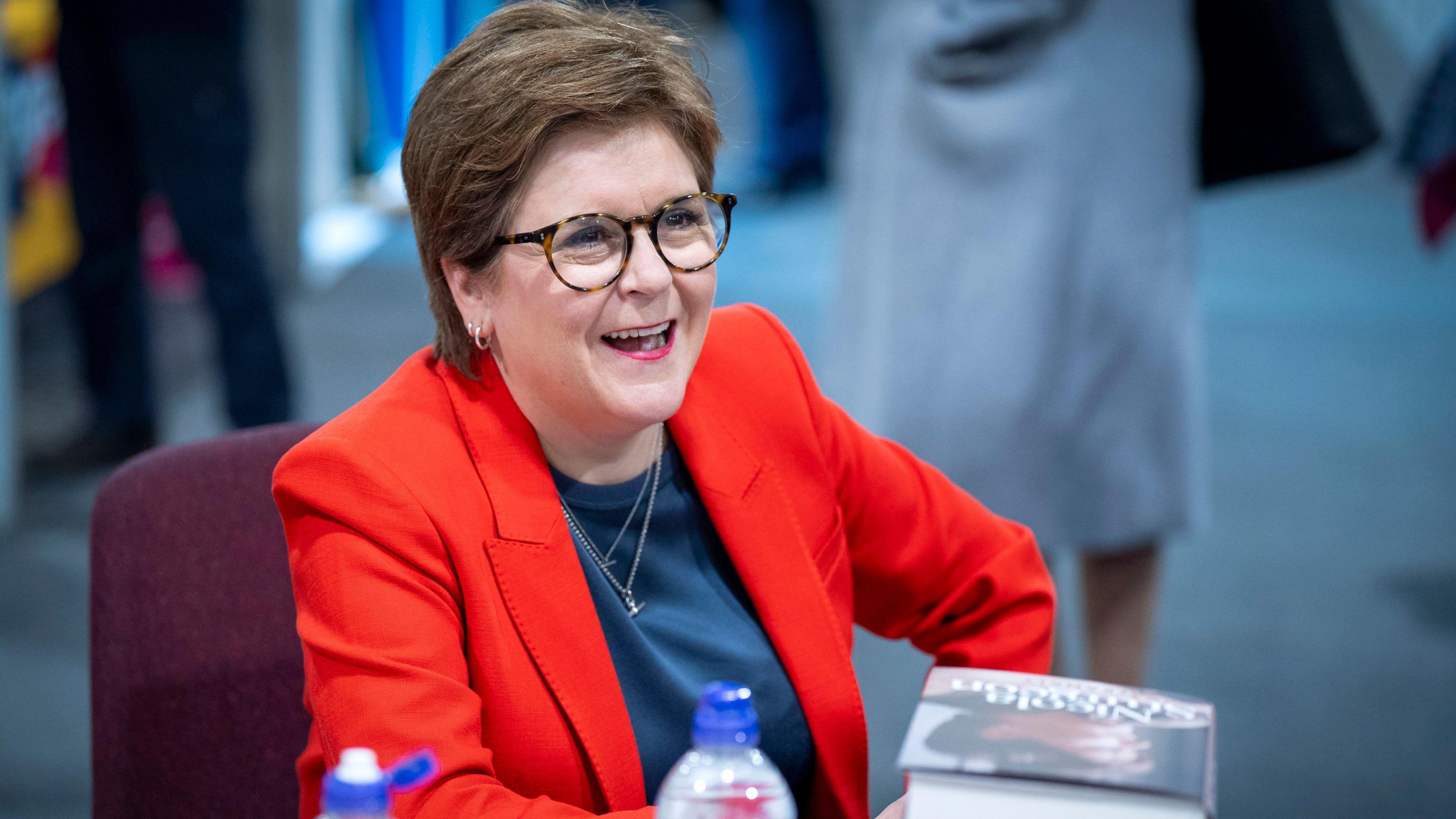 Former First Minister Nicola Sturgeon laughs as she signs copies of her memoir at the conference. She is wearing dark-rimmed glasses, a blue blouse and a bright red jacket.