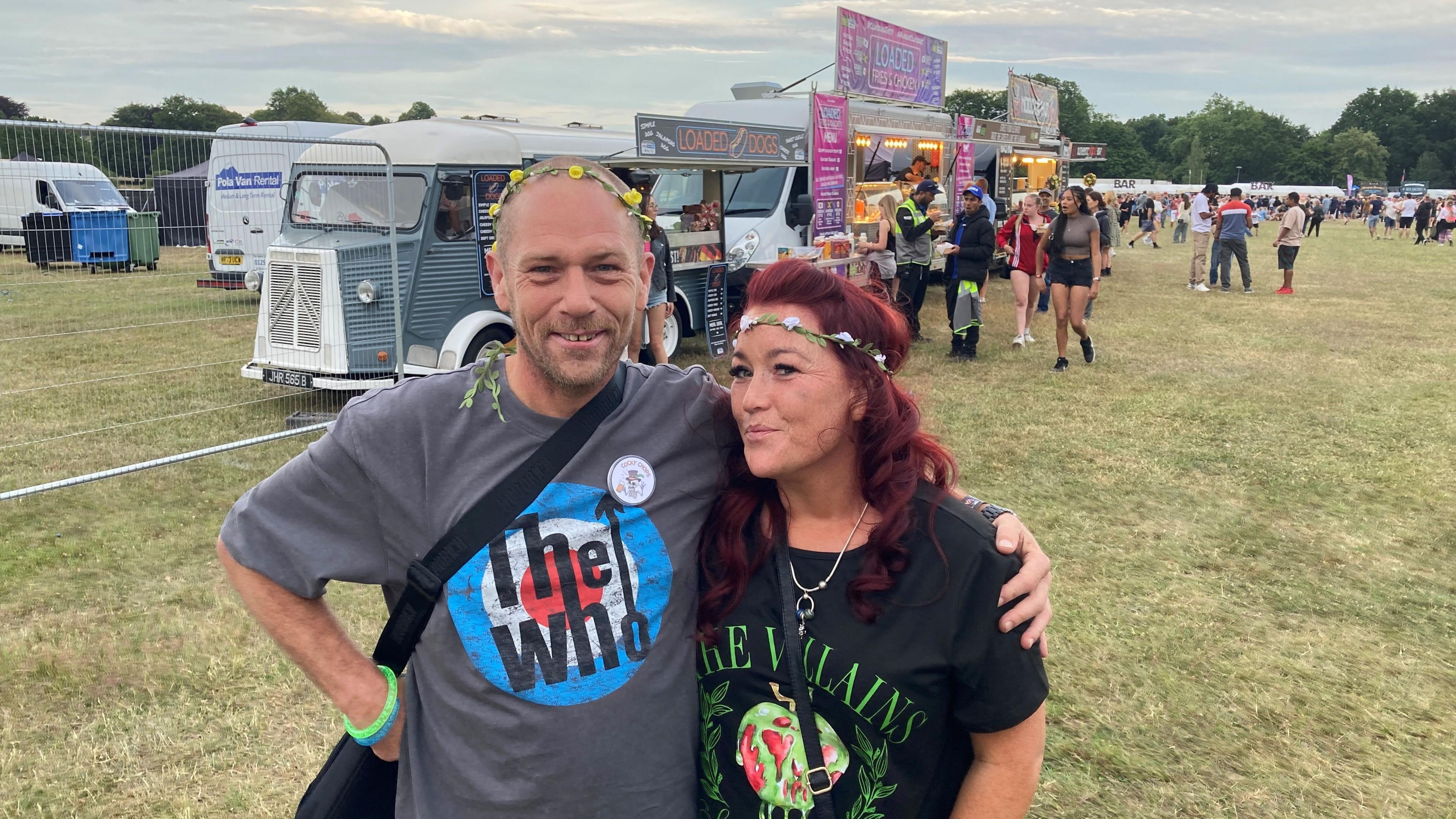 A man and woman stand in a field, with food stalls in the background. The man has a bald head and is wearing a T-shirt that says The Who on it. The woman has red hair and is wearing a T-shirt that says The Villains on it.
