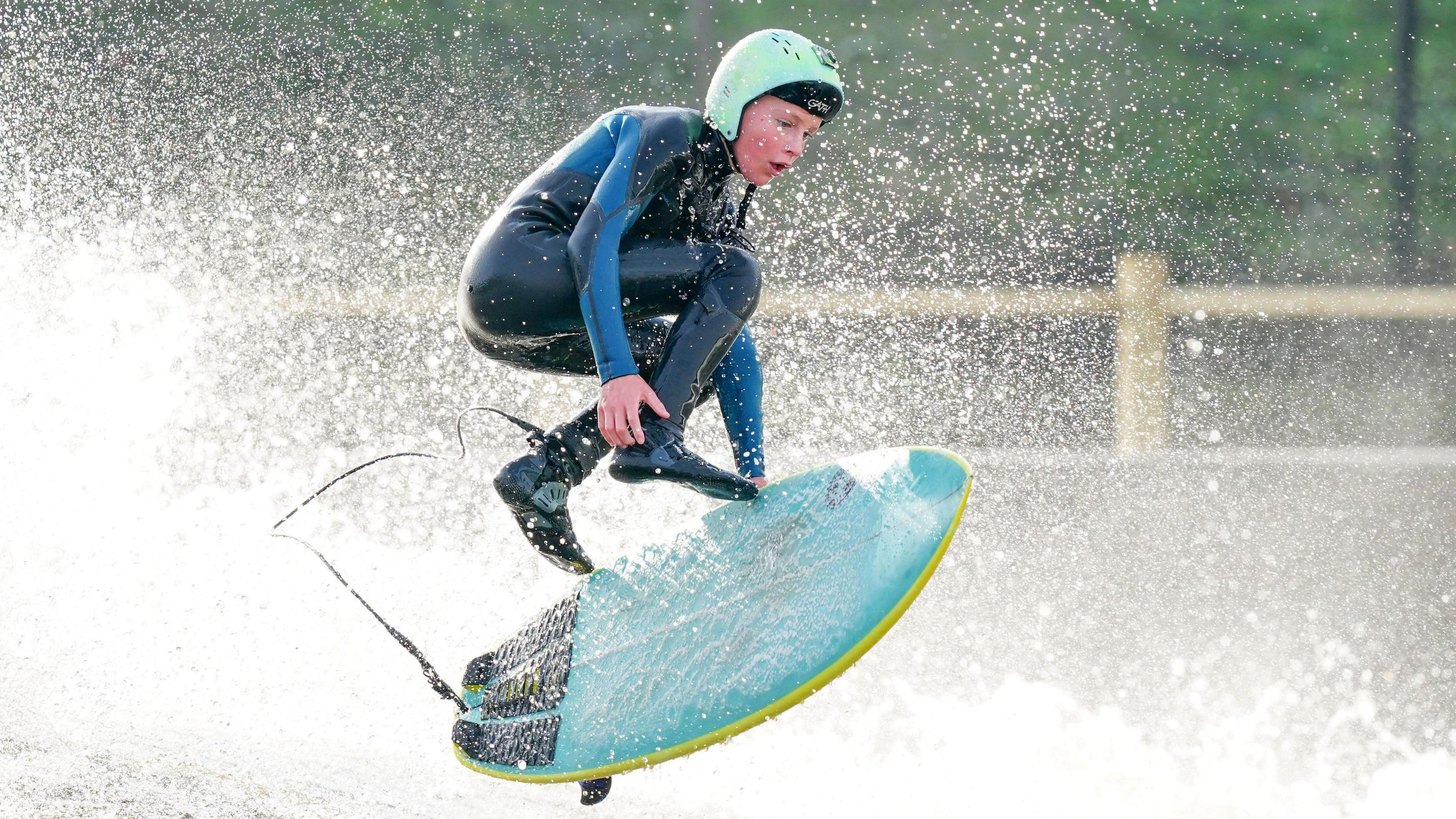 12 year old Kai jumps off his surfboard. He is holding onto a blue surfboard 