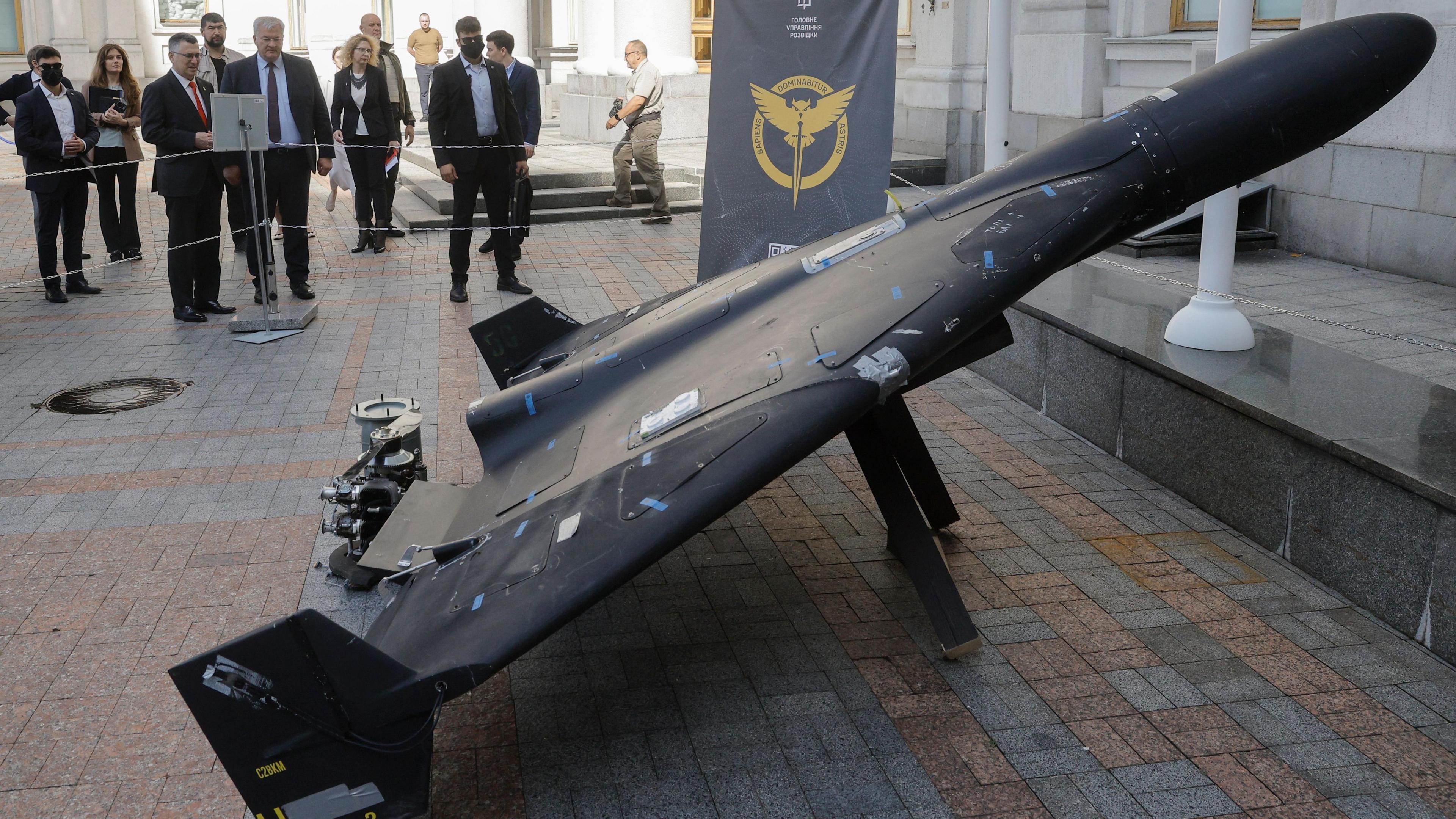 A captured Geran drone in Ukraine. It is shown in a square propped up against a stand. It appears to be lightly damaged and a crowd of people stand nearby examining it. A banner with the badge of Ukrainian defence intelligence can be seen in the background. 