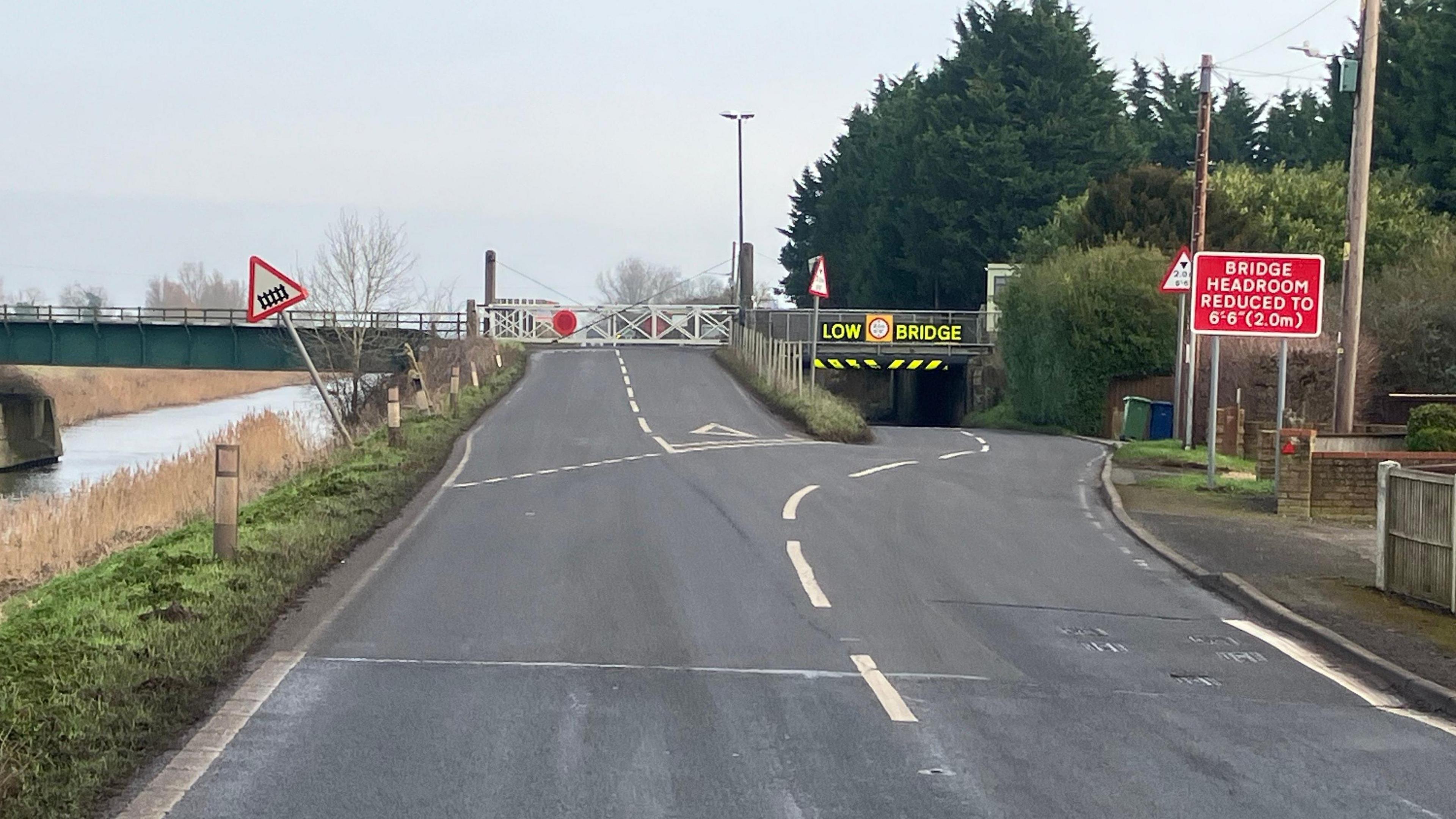 A low 2m (6 ft 6 inch) high underpass, with circular red and white warning signs that has a railway track above.
The is a yellow and black chevron that is damaged by vehicles hitting it.
There is a slight incline as the road goes up and goes over a manually operated gated section.