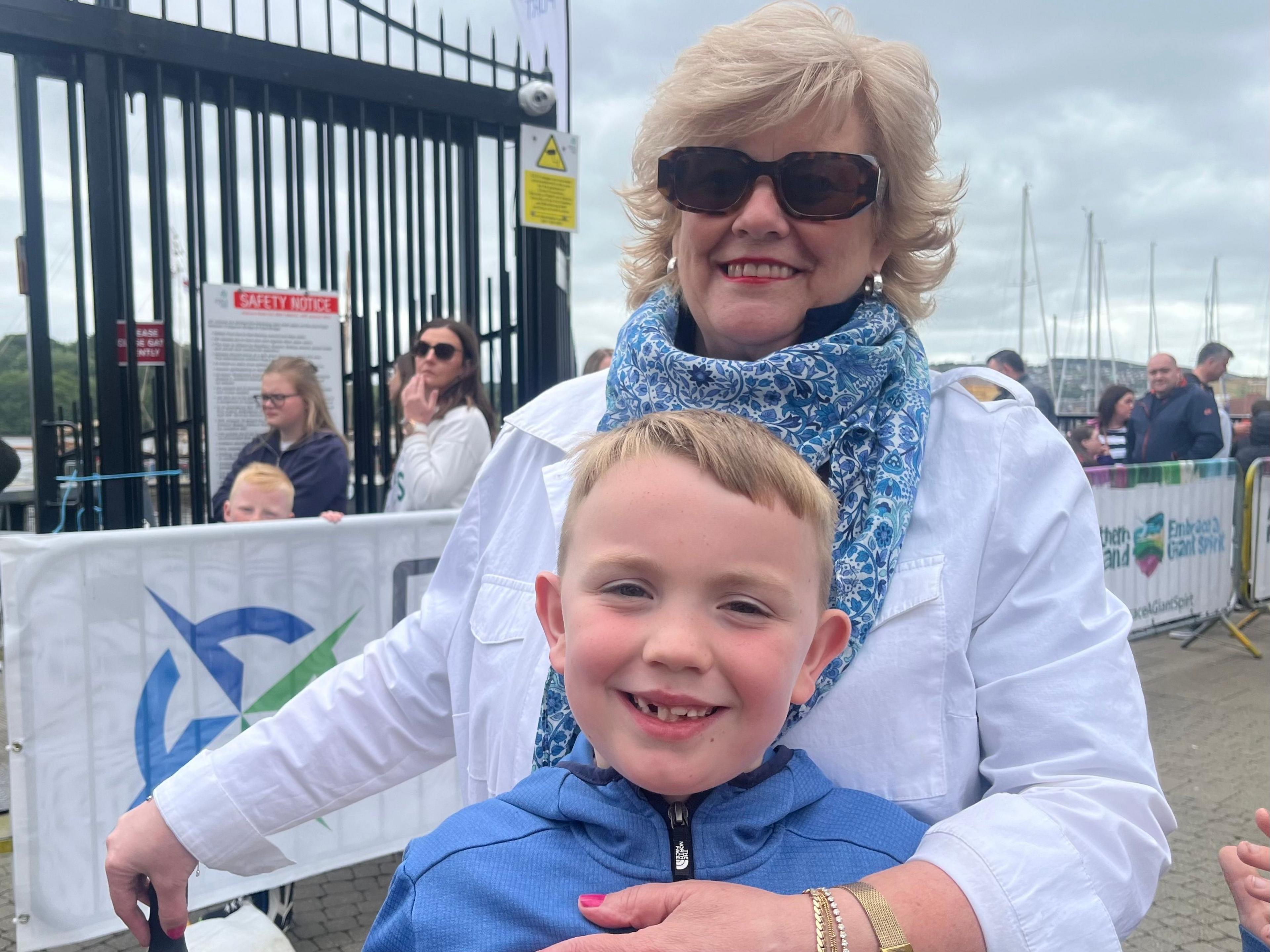 A woman, wearing sunglasses, standing in the middle of the photo, with a young boy with blonde hair standing in front of her 