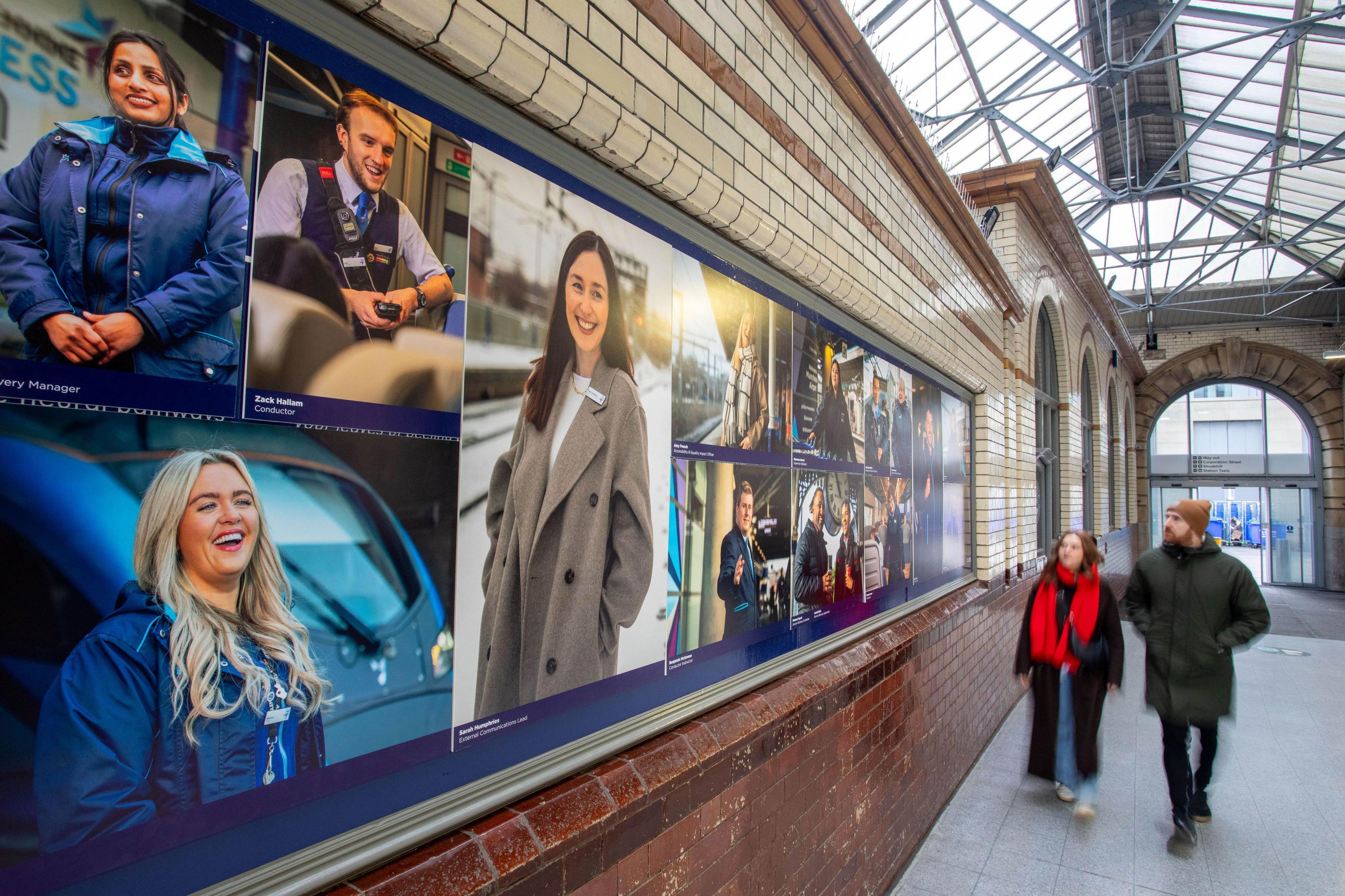 The exhibition in Manchester. It features a composite installation with multiple photographs of staff in a variety of locations. A man and a woman are walking past, looking at the pictures