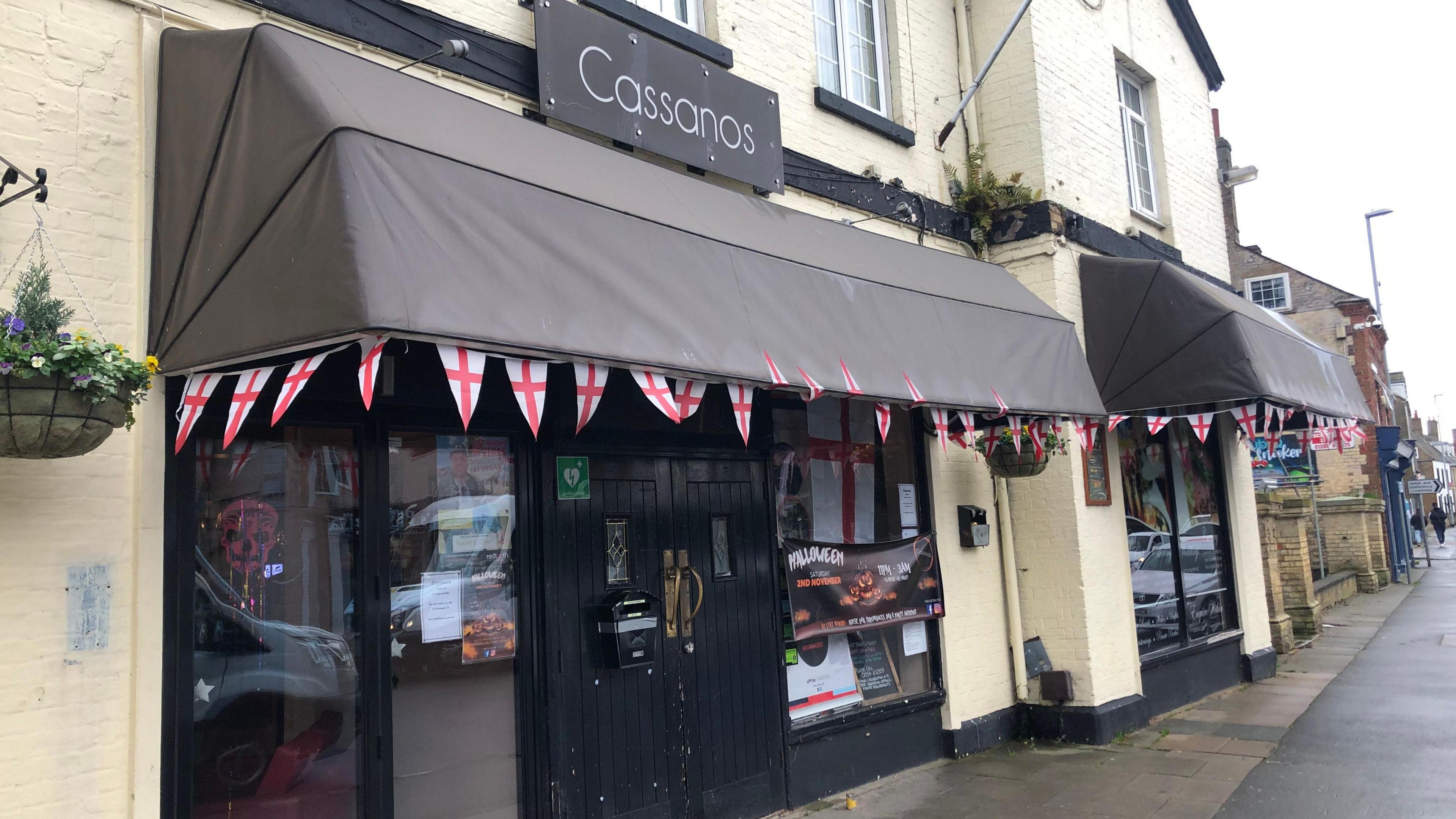 Pale yellow walls at the front entrance to 'Cassanos', some large windows can be seen at either end of the building and black wooden doors in the middle. There are two long, dark canopies over all windows and doors which has bunting in triangular design of the cross of St George fixed to it.