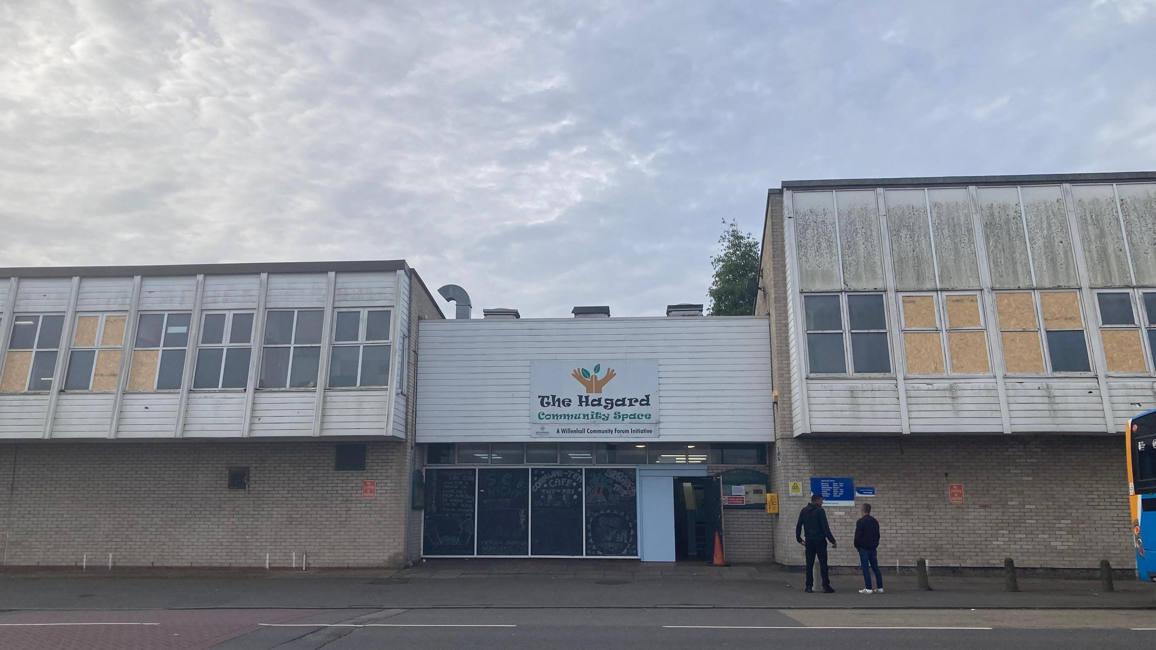 A community centre, with twp men standing in front of the building and a bus parked on the road outside.