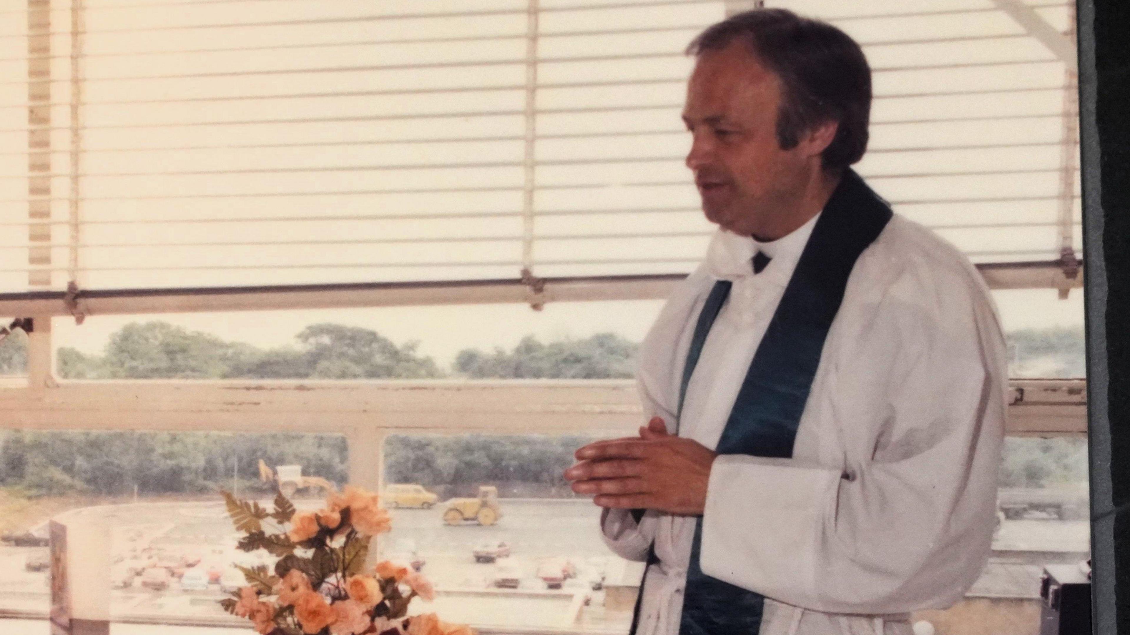 A younger Anthony Pierce stands clasping his hands with his back to a window. He is side of frame and is wearing white robes with a dark silk stole around his neck.