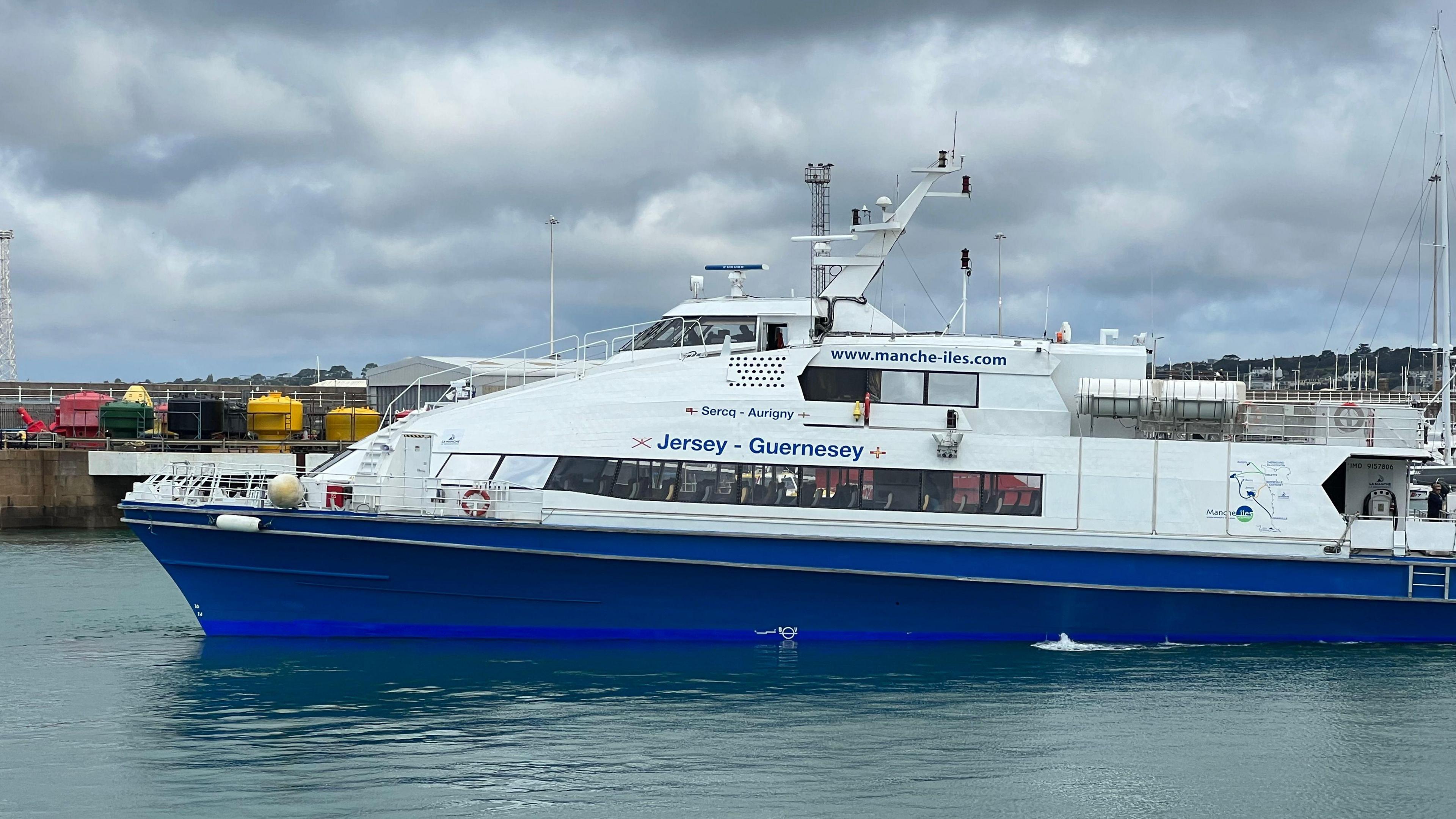 A passenger ferry with a blue hull and white top in a harbour near a granite pier.  It says www.mmanche-iles.com and has the names of the main Channel Islands in French: Sercq (Sark), Aurigny (Alderney), Jersey, Guernsey (Guernsey).