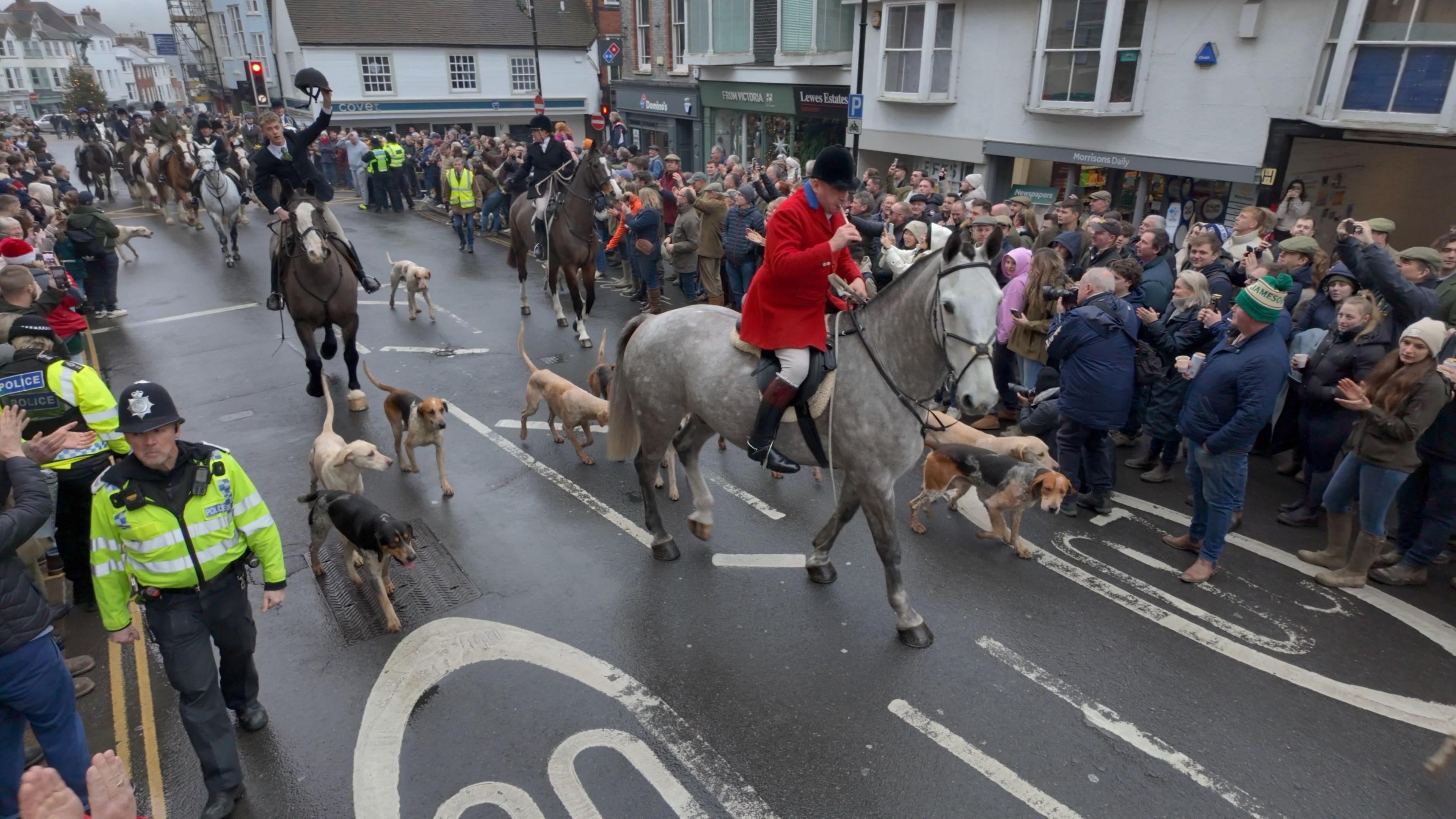 A grey horse ridden by a man in a red jacket leads other ridden horses through streets lines with police and protesters.