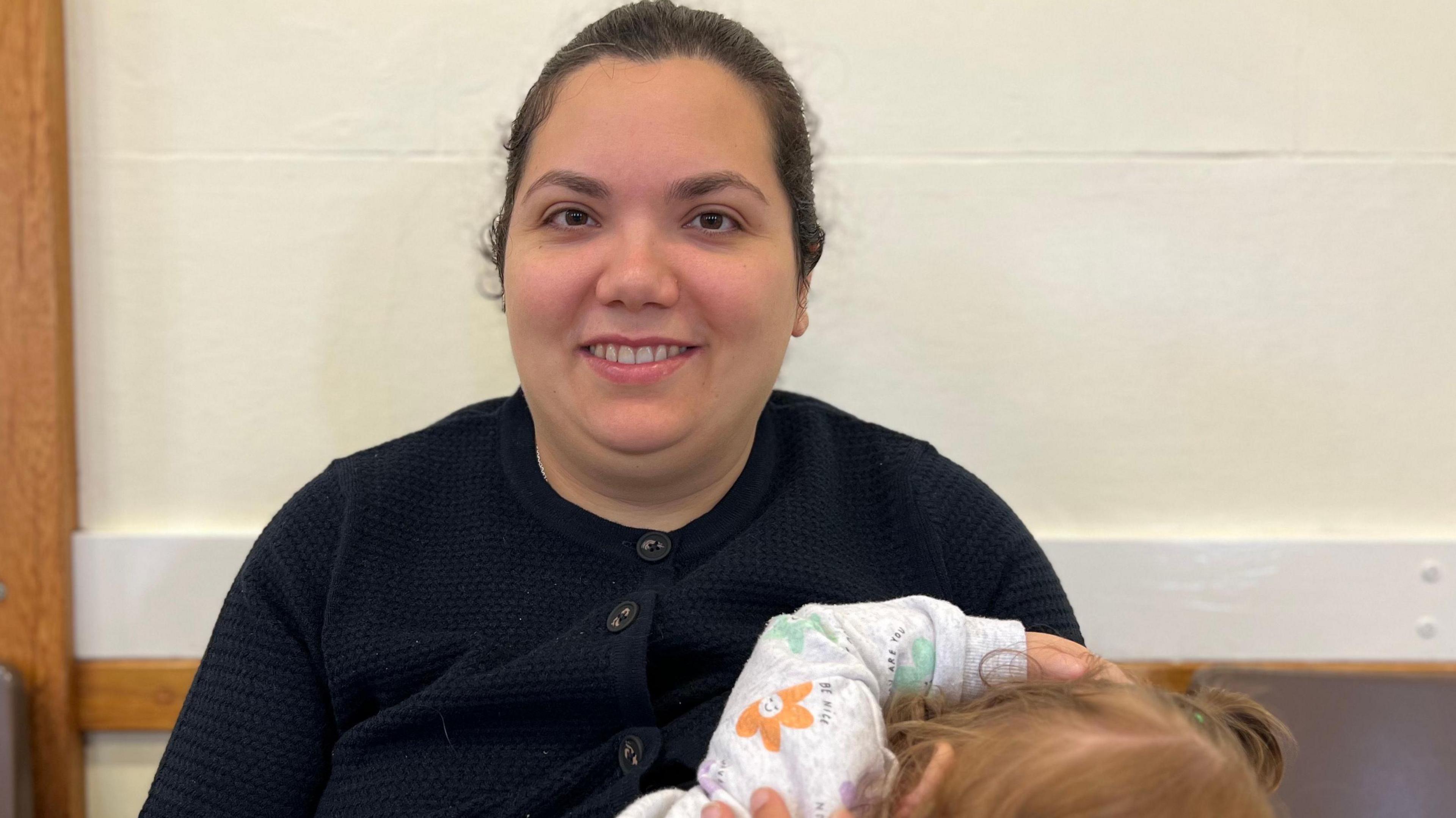 Stephanie Jackson pictured with dark brown hair tied back. She is wearing a black cardigan and holding her daughter.