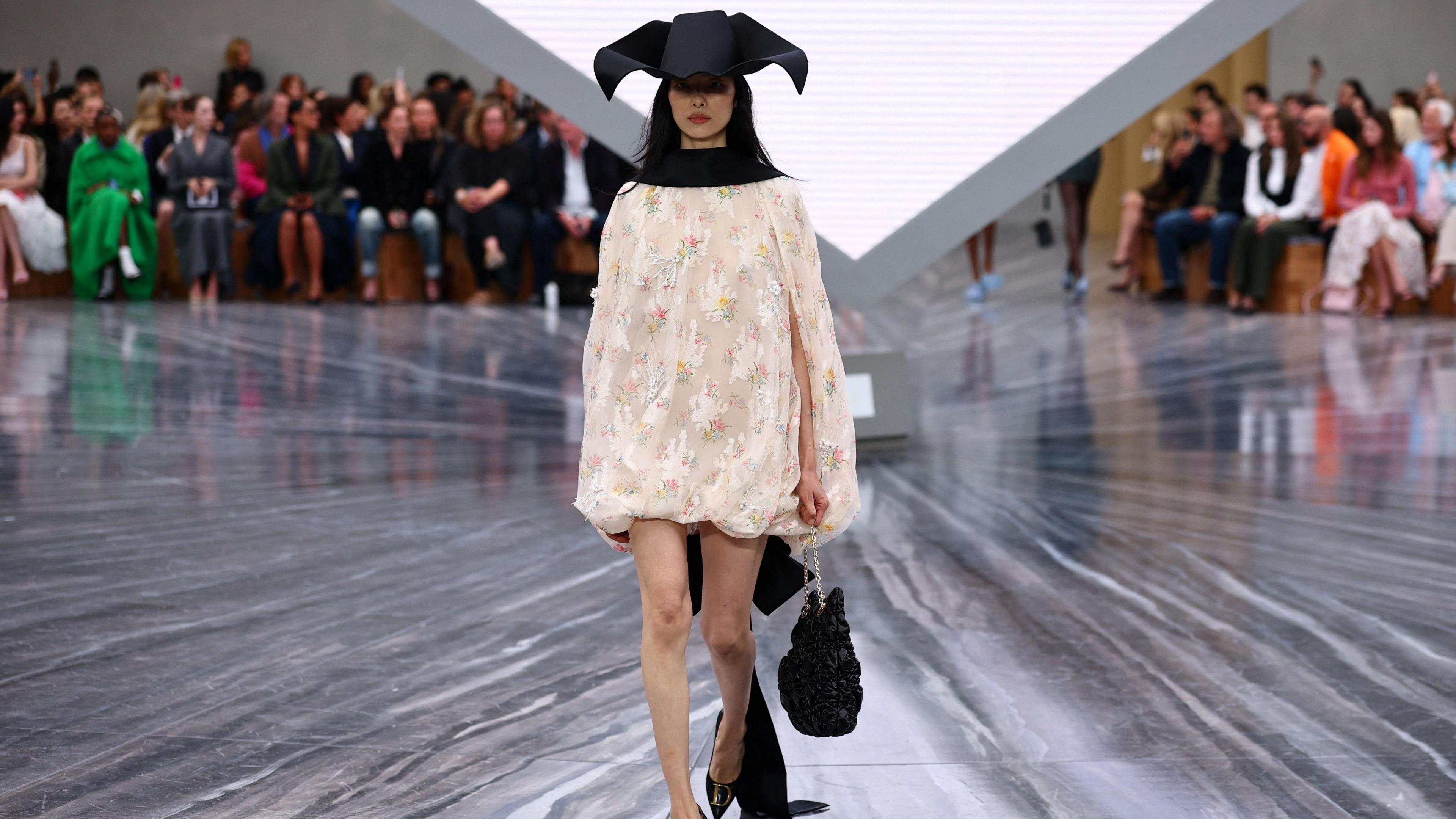 A woman in a cream floral dress with a black hat and black bag. She is on a catwalk, with the audience behind her.
