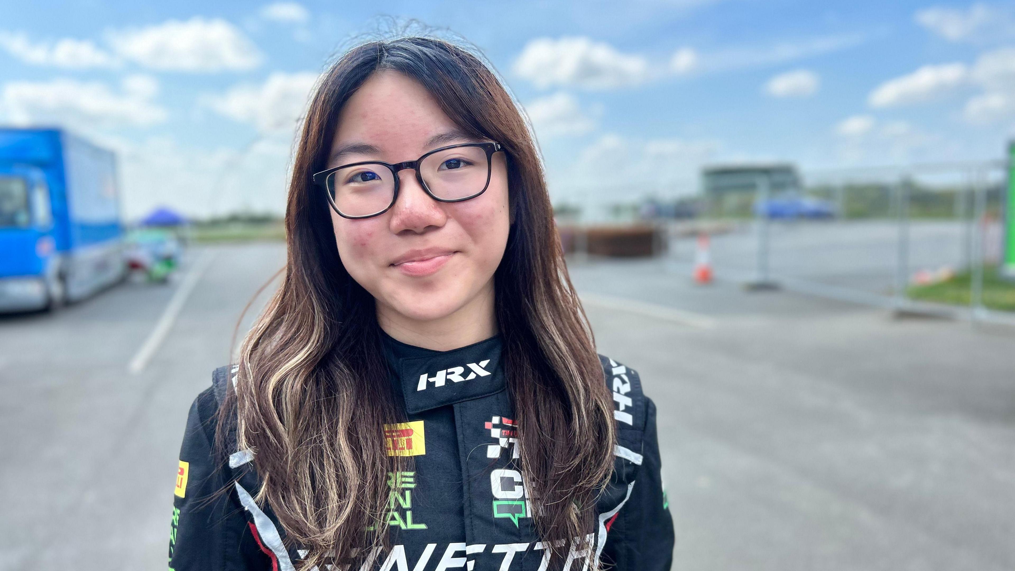 A girl with long hair wearing glasses smiling at the camera. She is wearing her racing suit standing behind the track