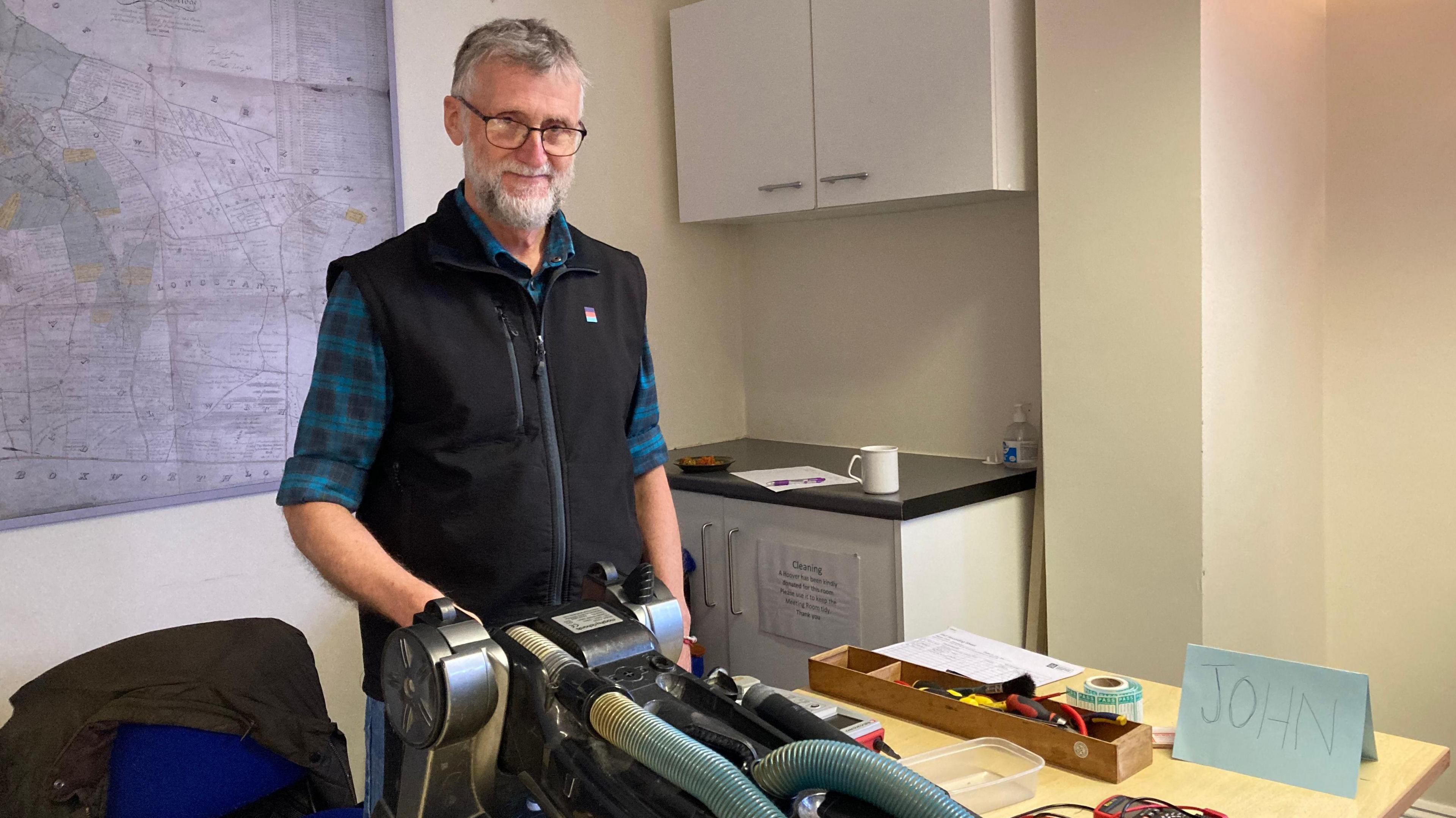 John Hallwood stands over a vacuum cleaner -he has a beard and is wearing a black gilet and checked shirt