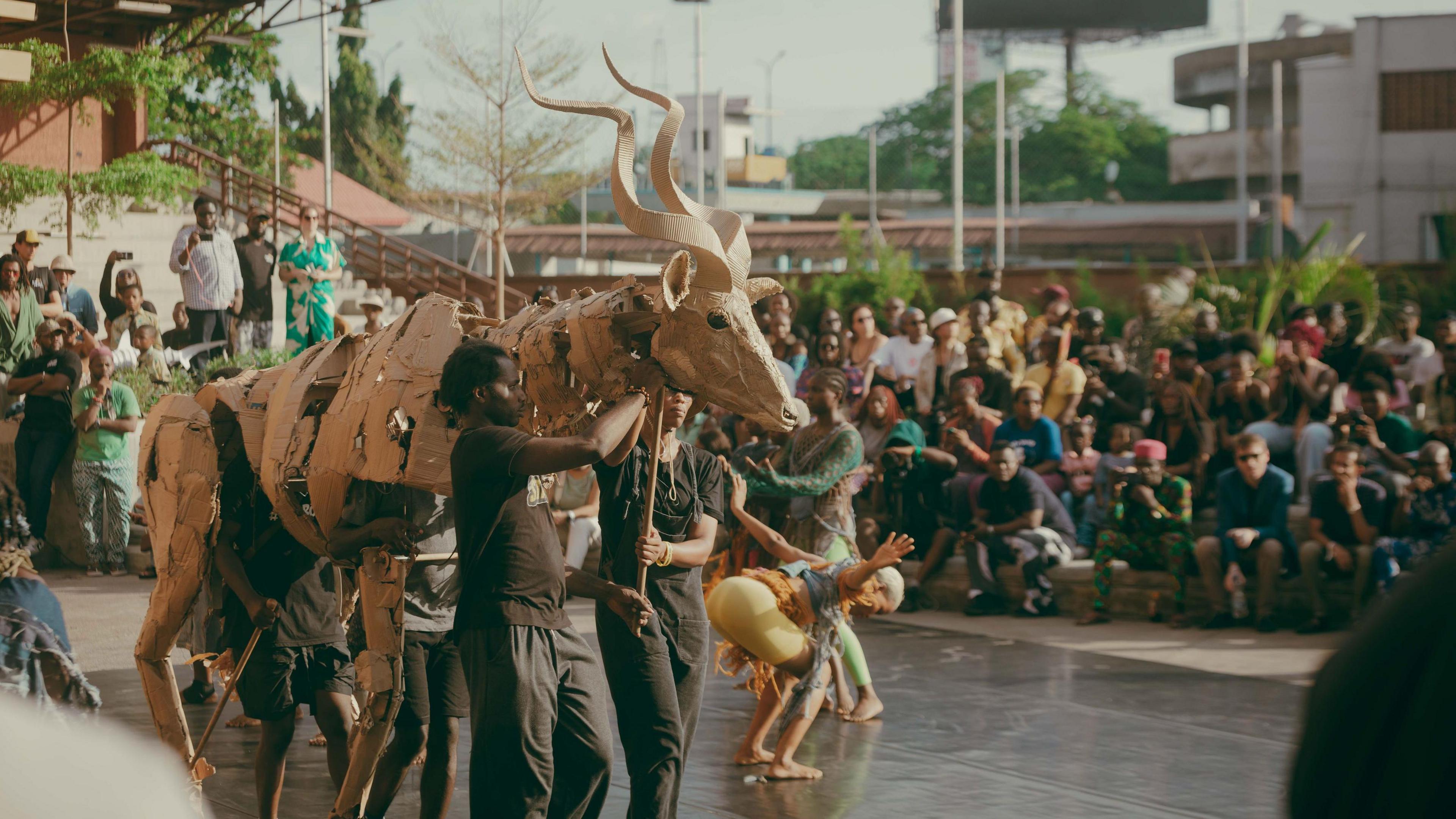 A large puppet animal with winding horns on a stage surrounded by performers with a large crowd of people watching in Lagos. 