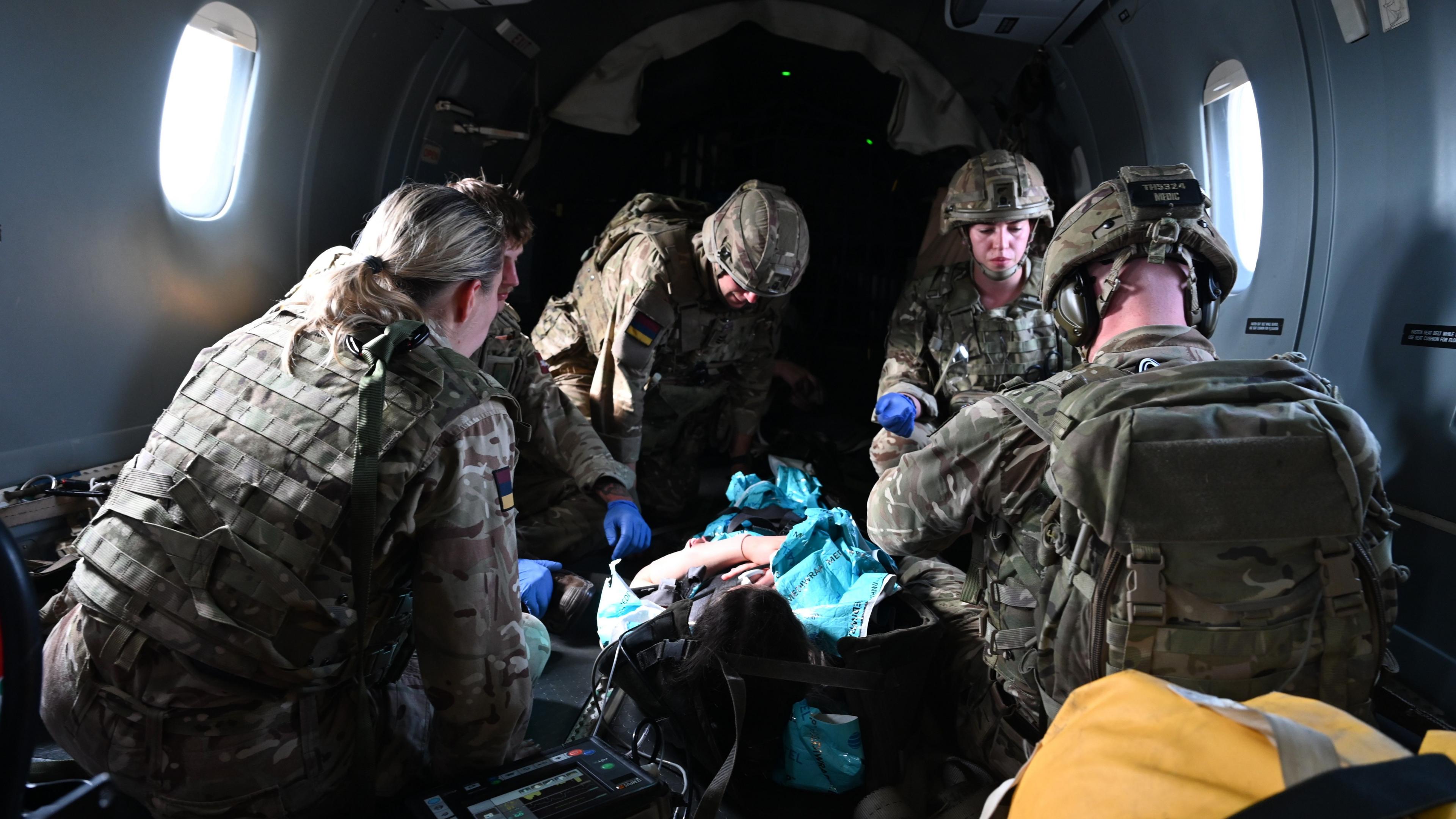 A medical simulation is carried out on-board an army aircraft. Military troops sit around a pretend casualty who lies on a stretched in the middle of the aircraft. 