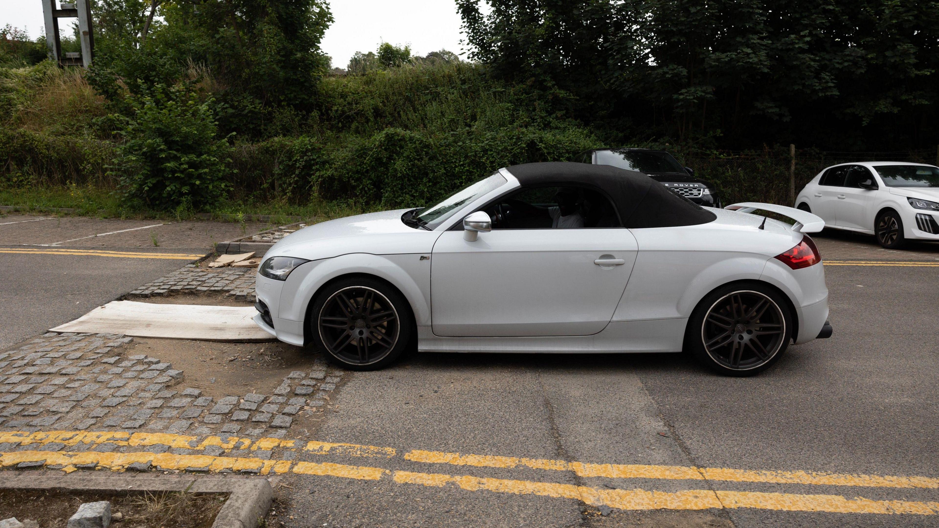 A white convertible car with a black soft top positioned just before the pothole/ broken speed bump. There are double yellow lines along both sides of the road. There’s another white car parked in the background, and there are green trees and bushes.