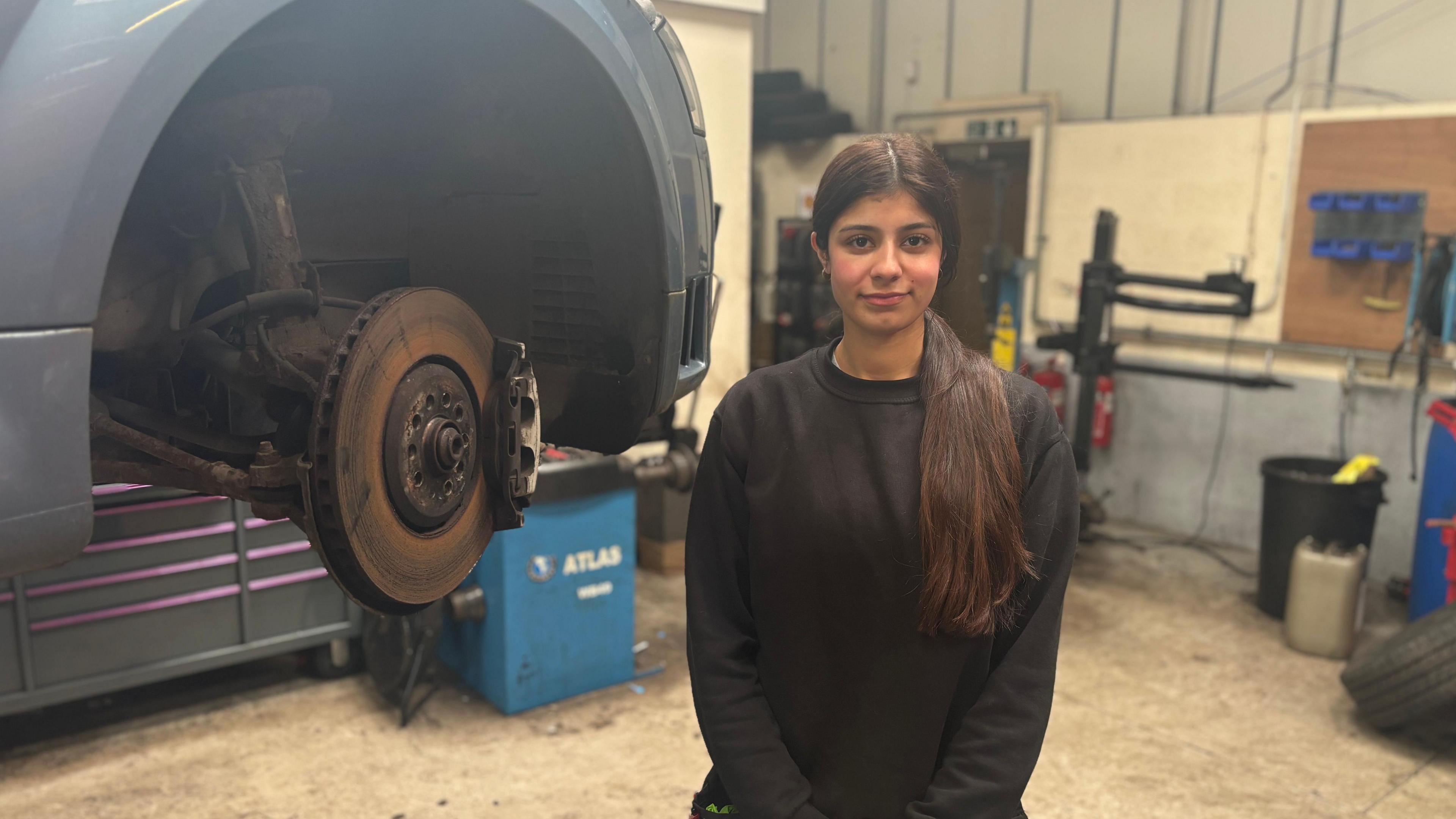 Sophie stands in front of a stripped wheel arch. She has long brown hair pulled over her shoulder in a ponytail. She wears a black jumper.