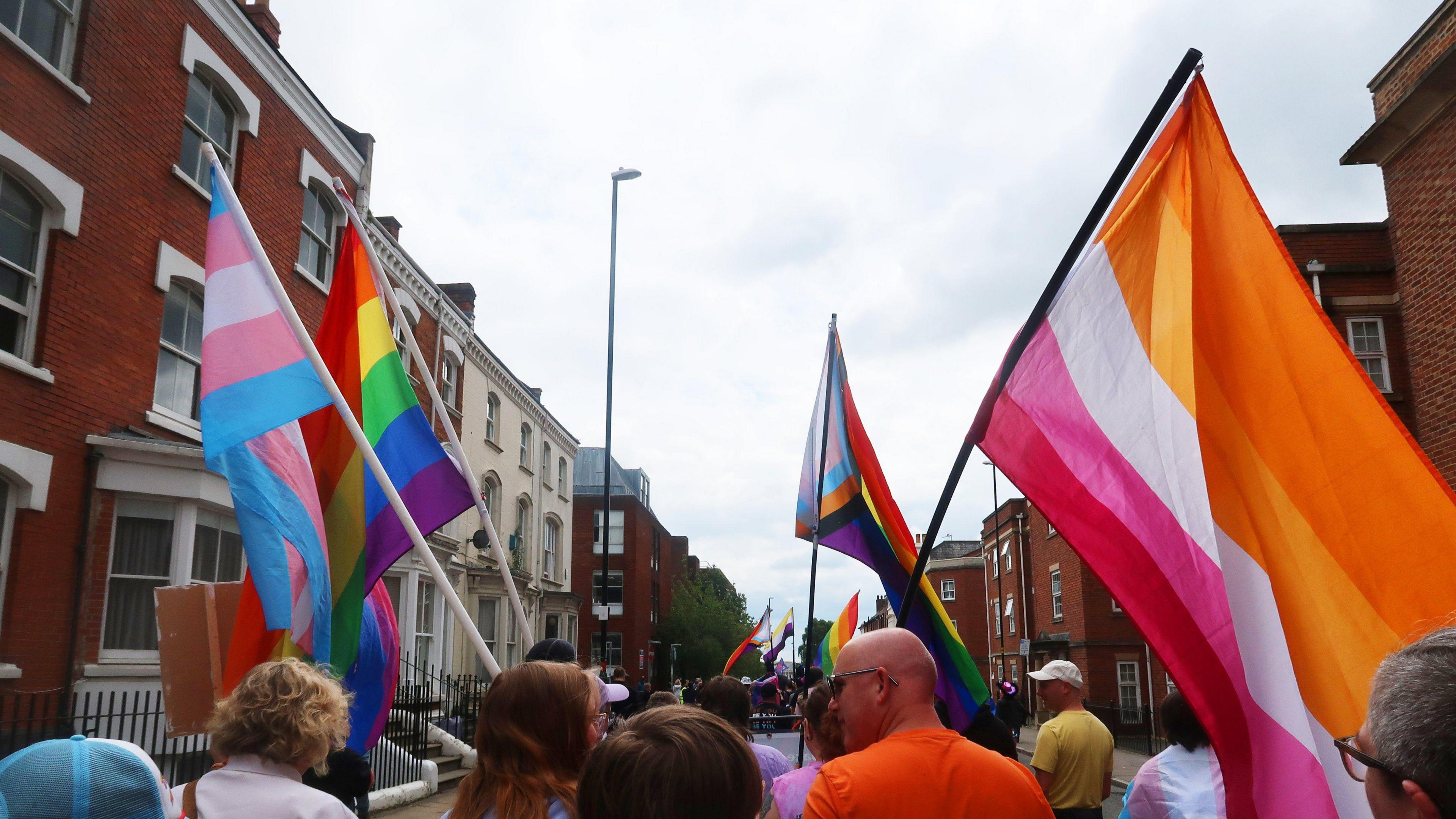 Pride flags and other colourful flags held aloft at the head of a parade passed terraced houses and businesses