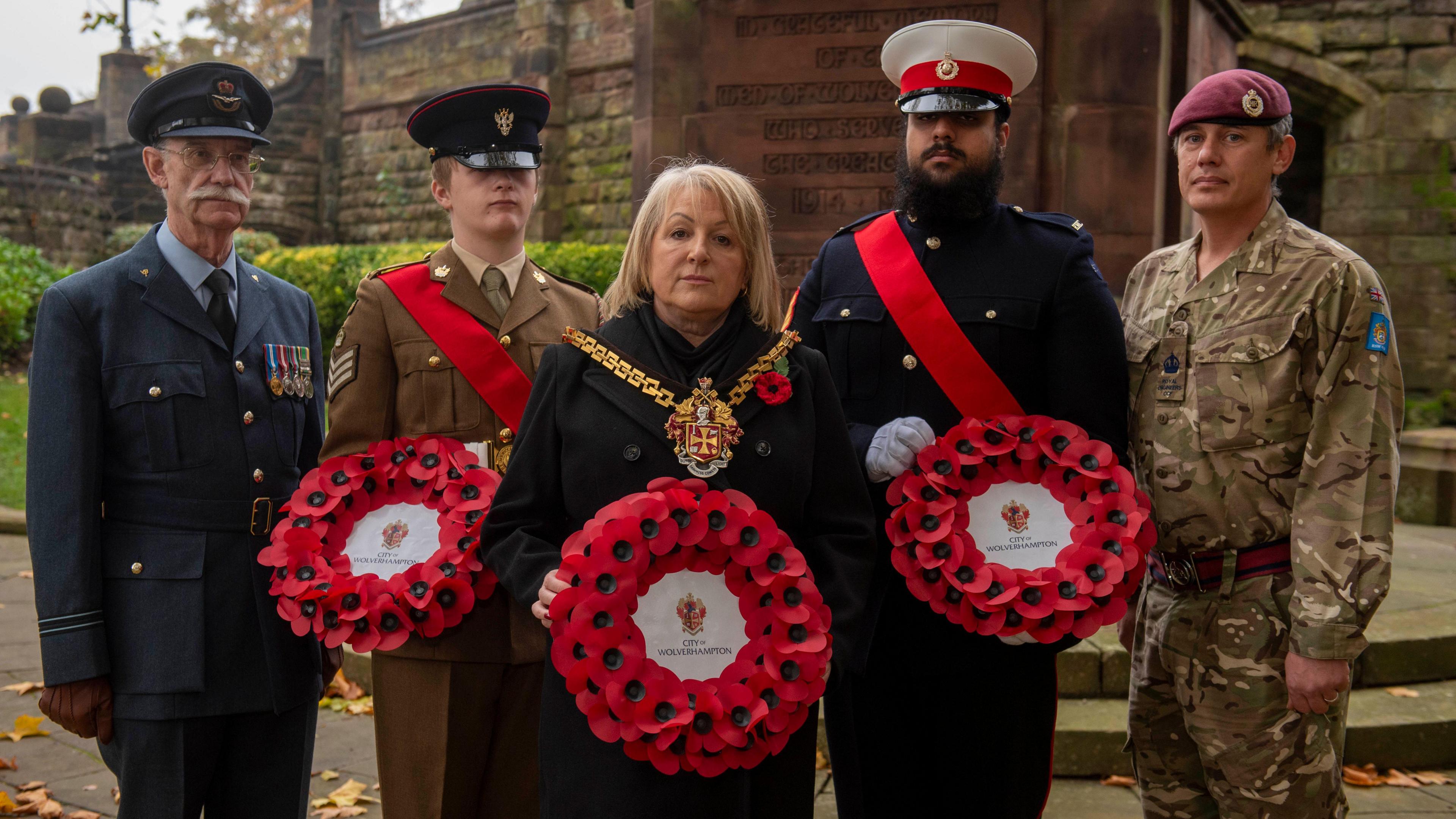 Linda Leach standing wearing a mayoral chain holding a poppy wreath. She is with four men in different armed forces uniforms wearing medals and also holding poppy wreaths