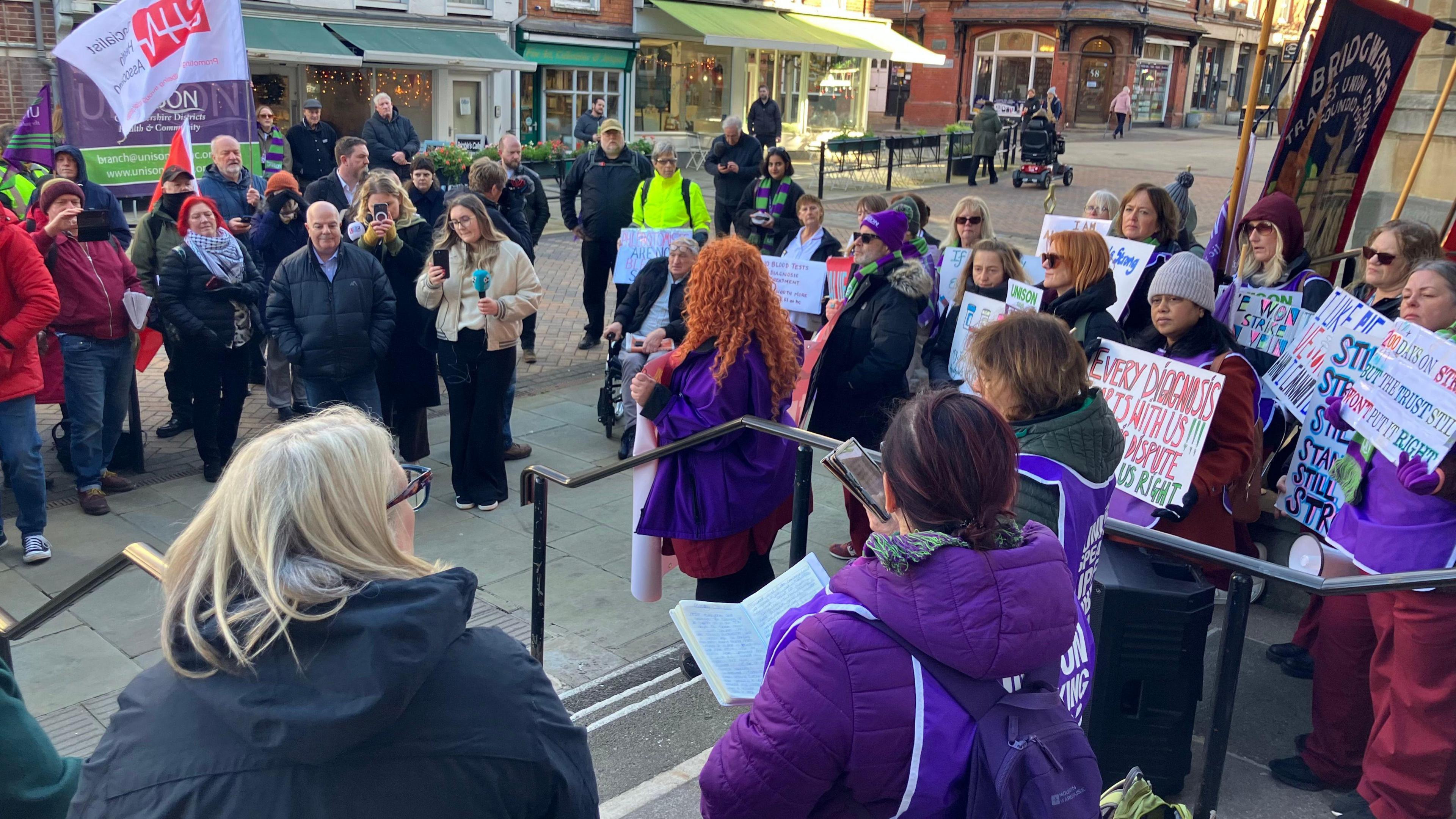 A crowd of people gather as part of a rally in Gloucester city centre, outside Shire Hall, with placards and purple clothing. A woman with curly, bright ginger hair makes a speech into a microphone on the stairs.