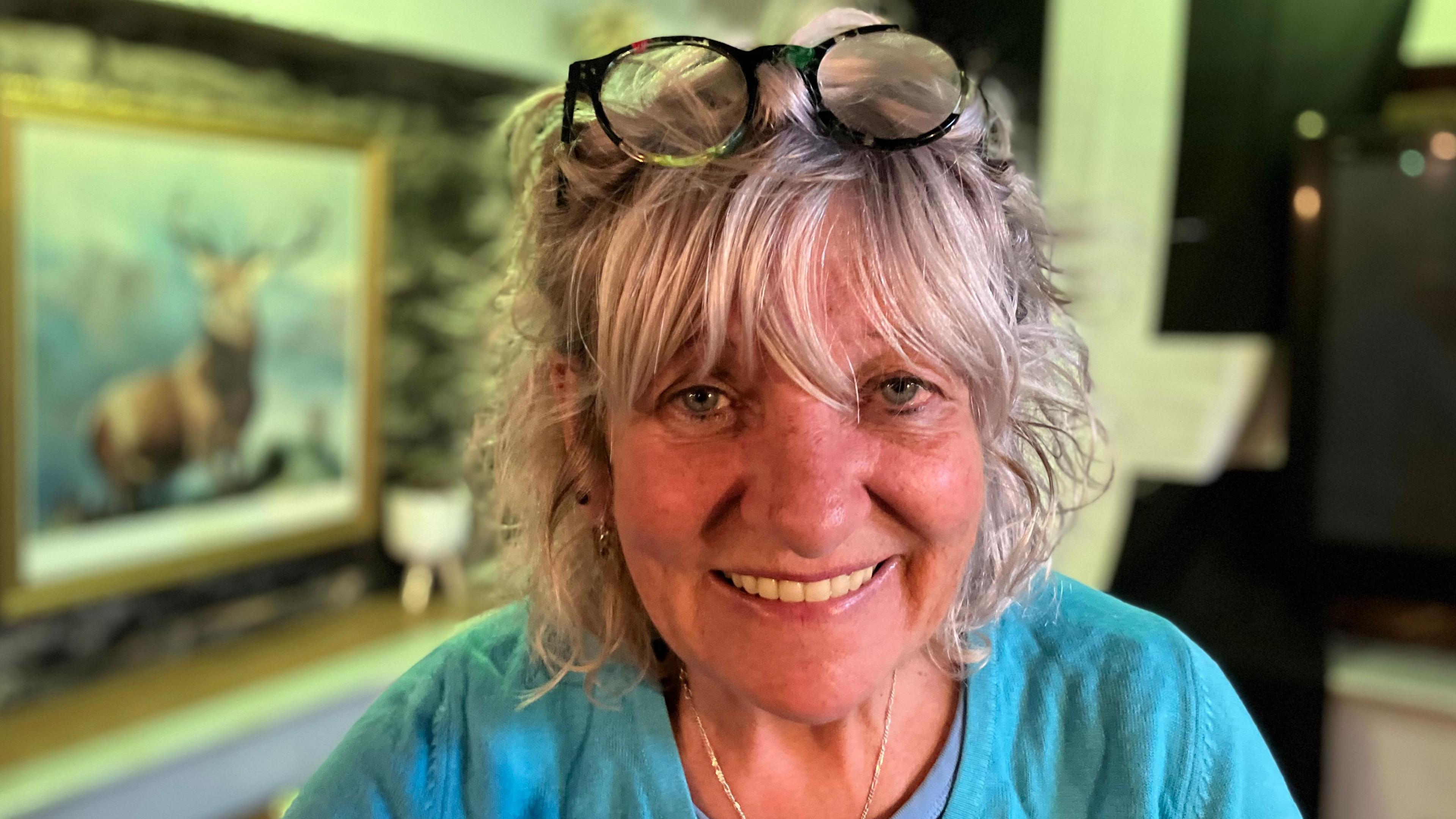 Trina Crawford smiling at the camera. She is wearing a blue t-shirt and has a pair of black glasses on top of her head. In the background is the pub, and a large painting of a deer on the left.