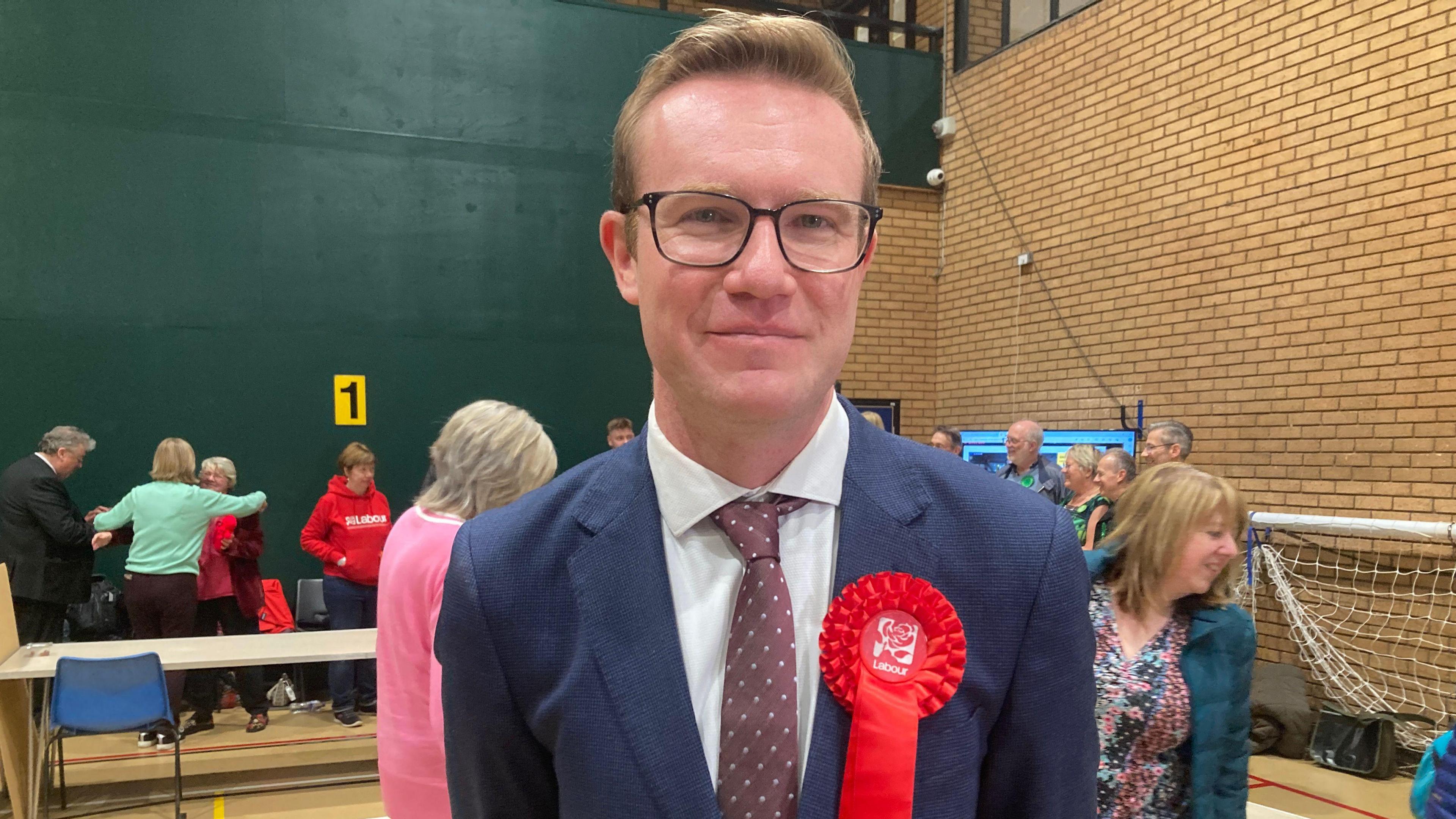 Tim Roca standing in a community sports hall after being elected Labour MP. He is wearing glasses, a navy blue suit and red tie with white spots, and has a red Labour rosette pinned to his lapel.