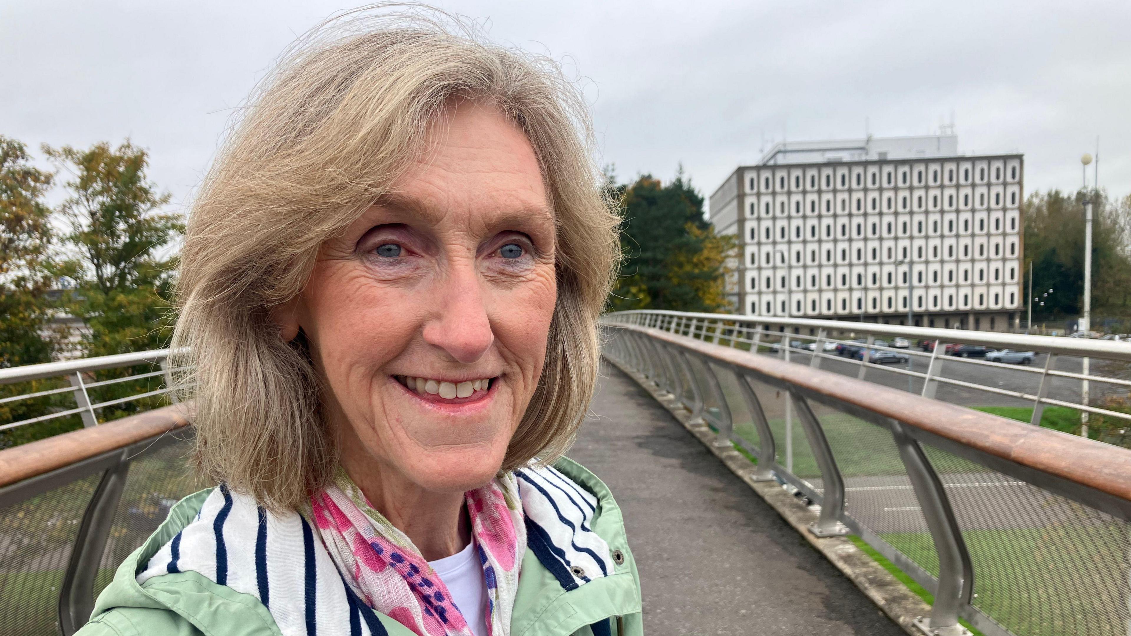 Maureen Millar pictured at a bridge leading to Marlborough House. She has shoulder length light hair and is wearing a green coat with a pink patterned scarf. She is smiling.
