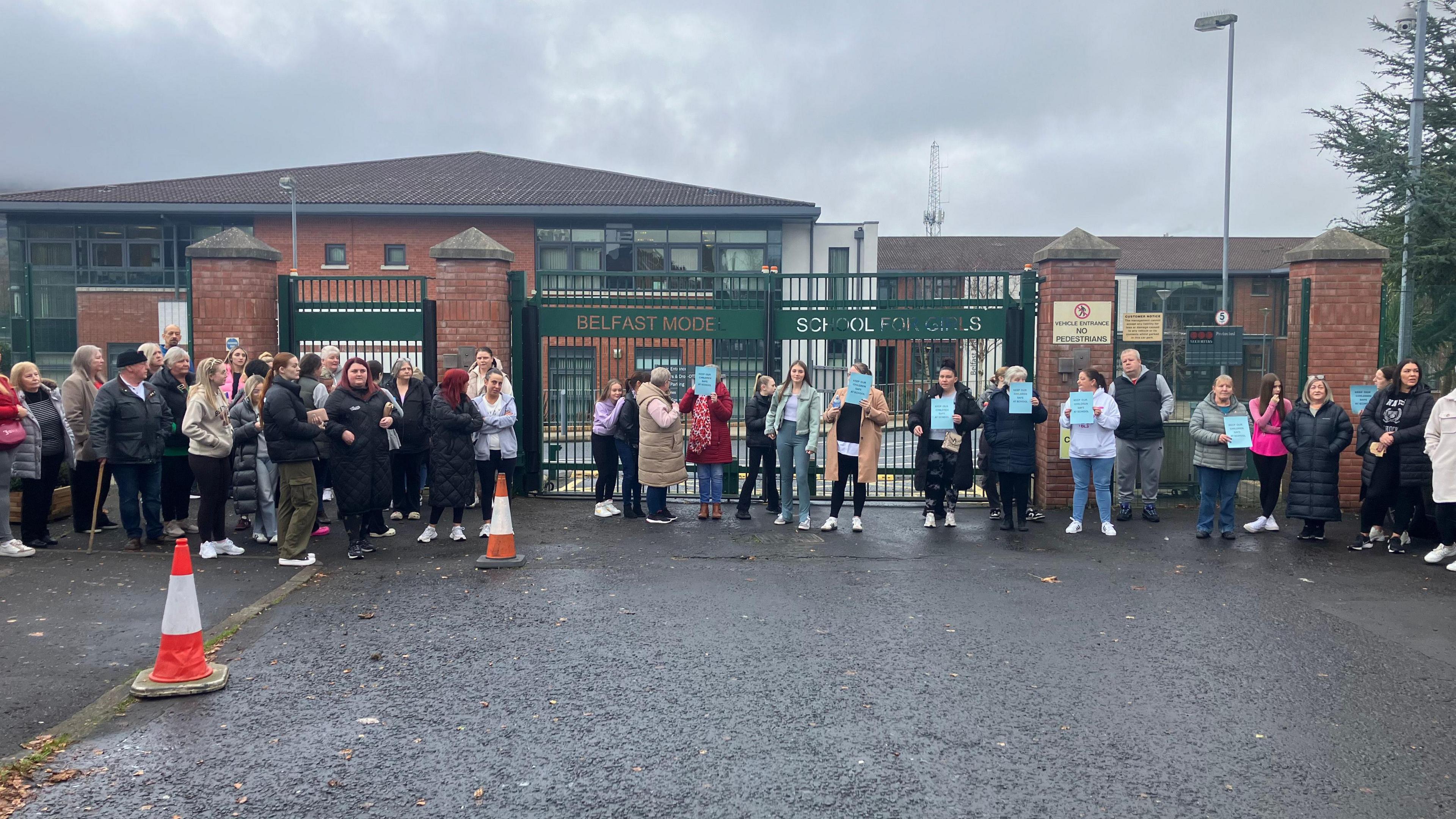 Dozens of people standing outside school gates. They are holding up blue placards but the writing cannot be made out. The gates are green wrought iron and the school behind them is a modern red-brick building.