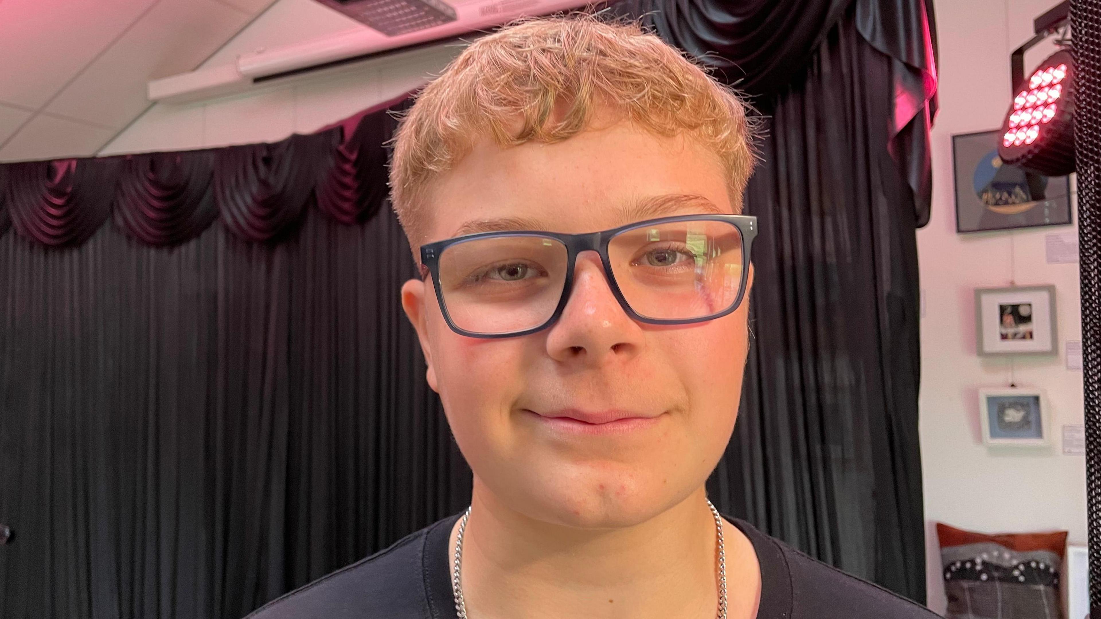 A teenage boy with short blonde curly hair and black-rimmed glasses stands in front of a black backdrop.