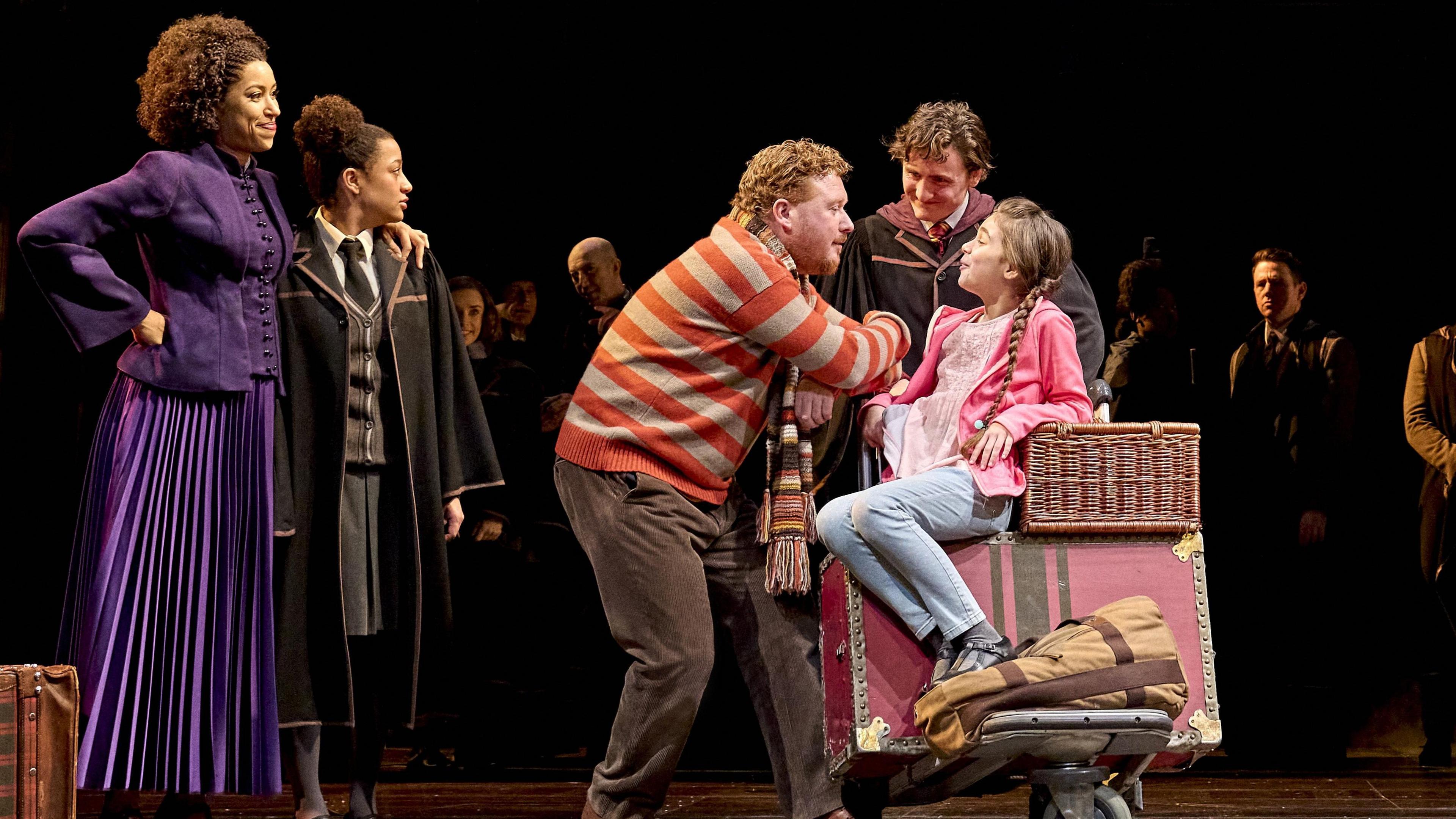 The cast from the play Harry Potter and the Cursed Child on stage.
There is a girl sitting on a suitcase with a man playing her dad beside her
Behind her is a woman wearing a purple gown standing next to a teenager.
There are a few people standing behind them too.
