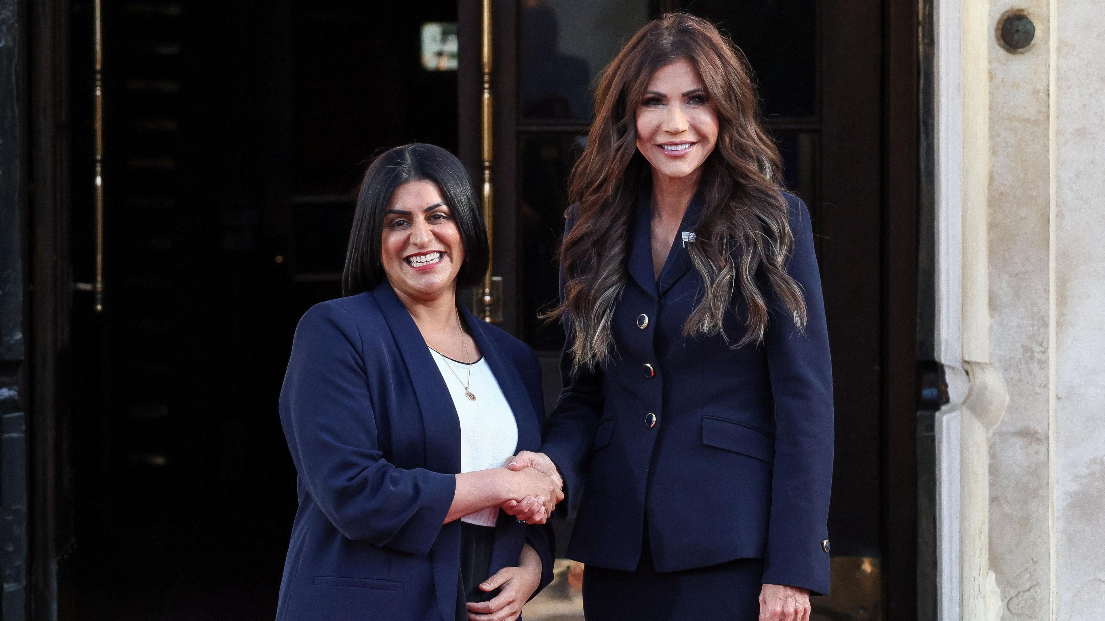 Home Secretary Shabana Mahmood shakes hands with U.S. Homeland Security Secretary Kristi Noem . Mahmood and Noem are both smiling and wearing navy blue suit jackets.