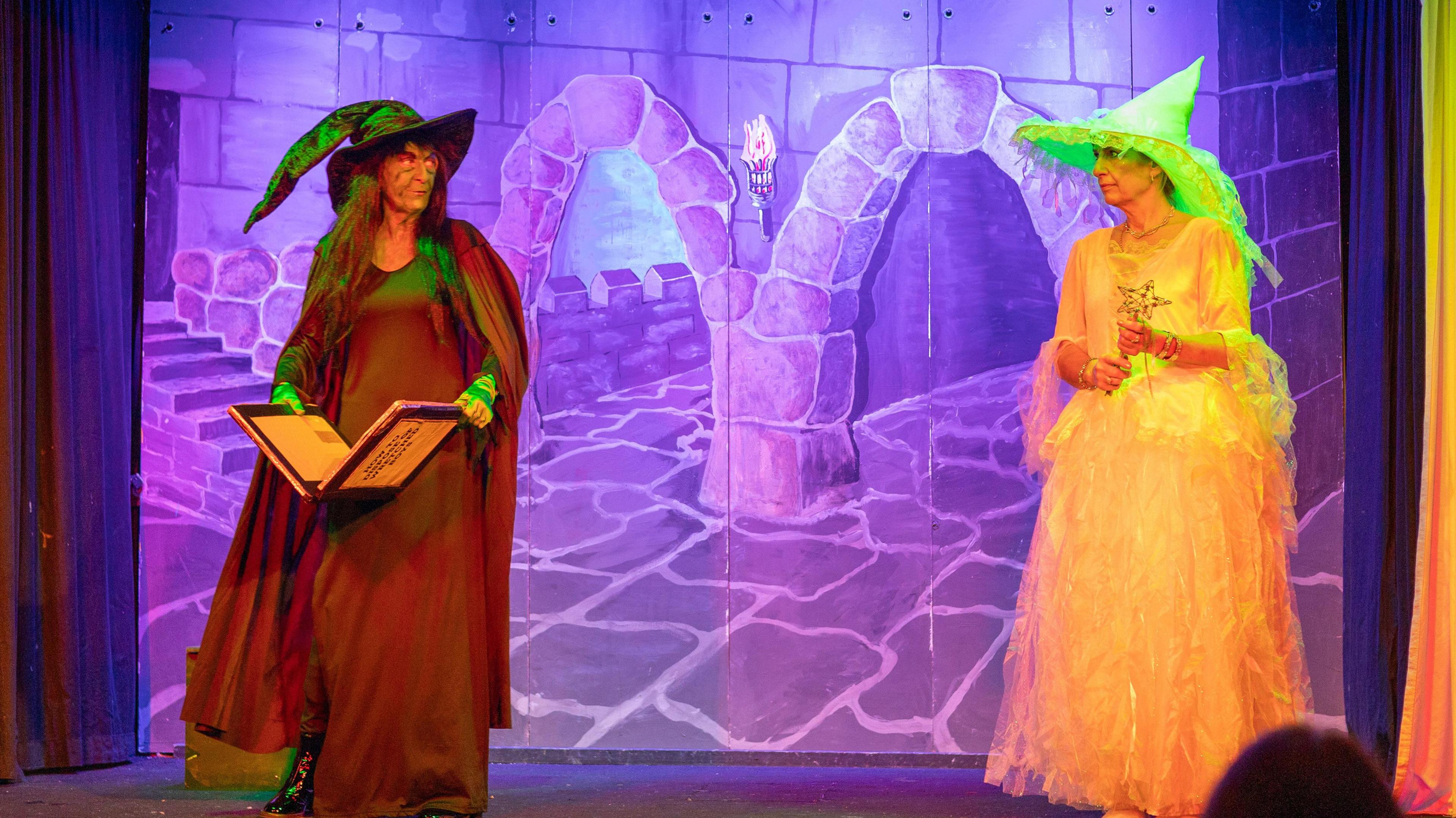 Two women on stage mid-way through a scene, with a purple backdrop of a brick road. One woman is dressed as the Wicked Witch of the West from Wizard of Oz, with a book in her hands, and the other is dressed as Glinda the Good Witch.