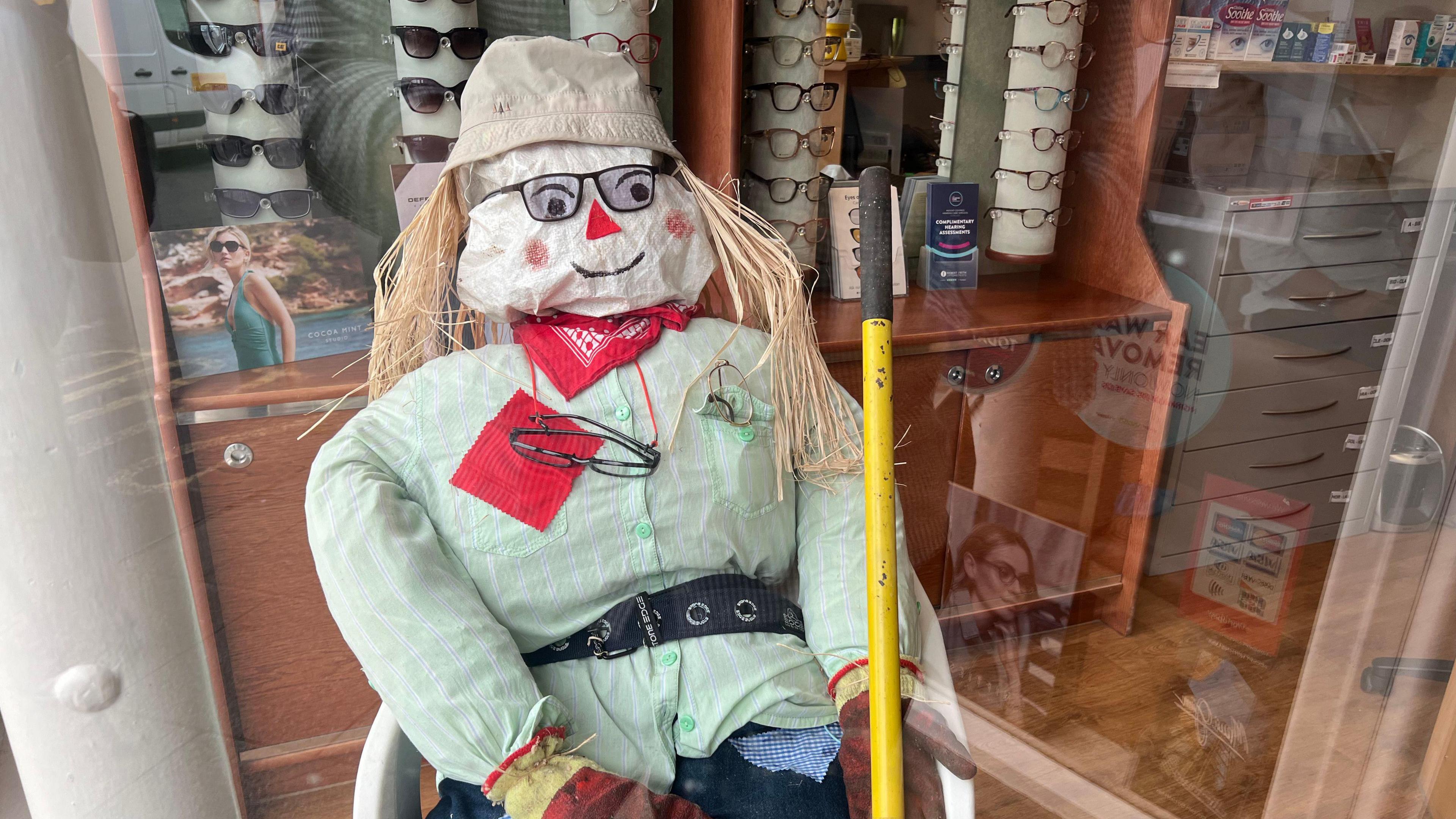 A traditional looking scarecrow sits in the window of a opticians, wearing glasses. He is surrounded by racks of glasses. 