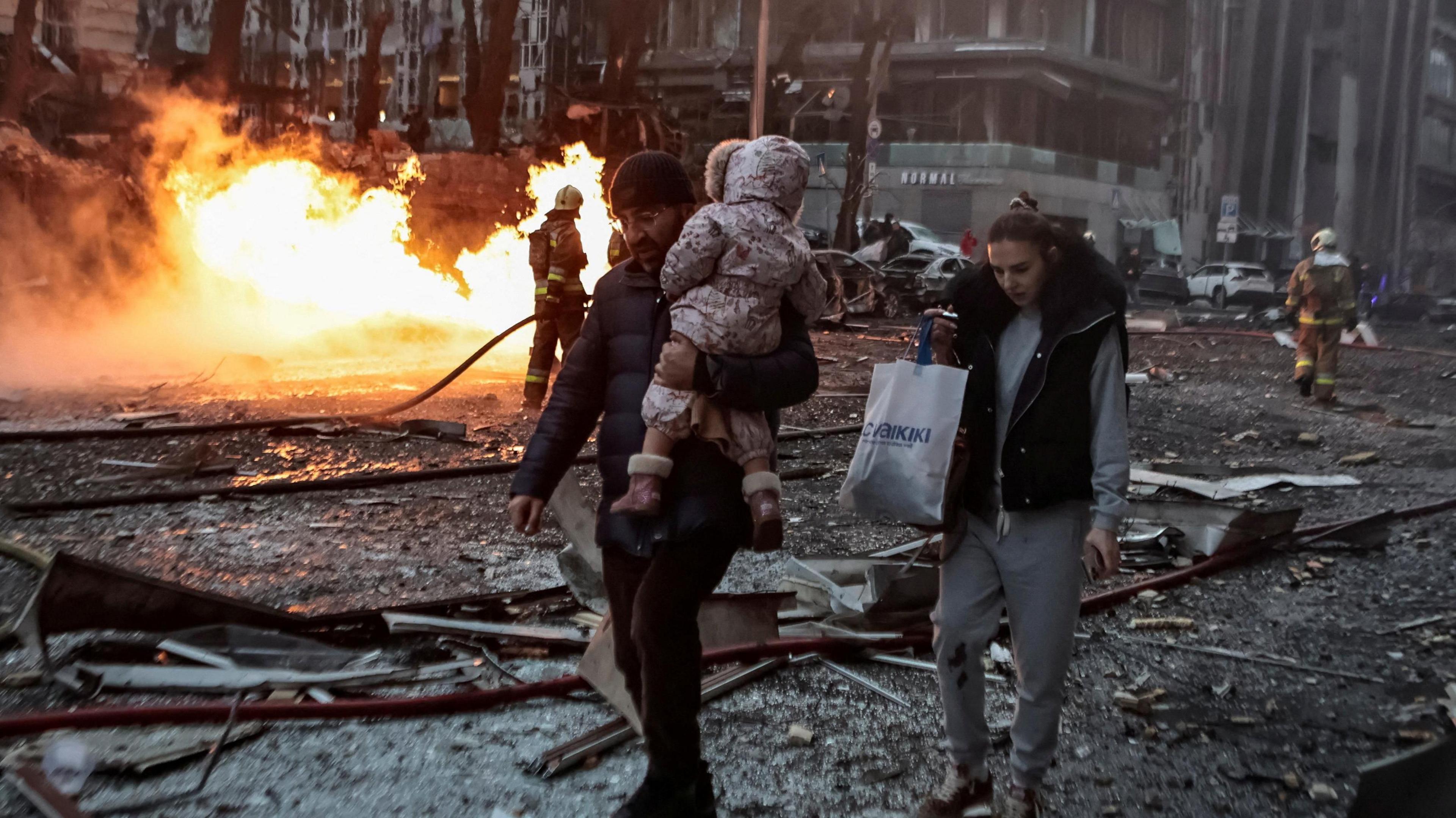 A man holding a baby walking away with flames and debris on a road 