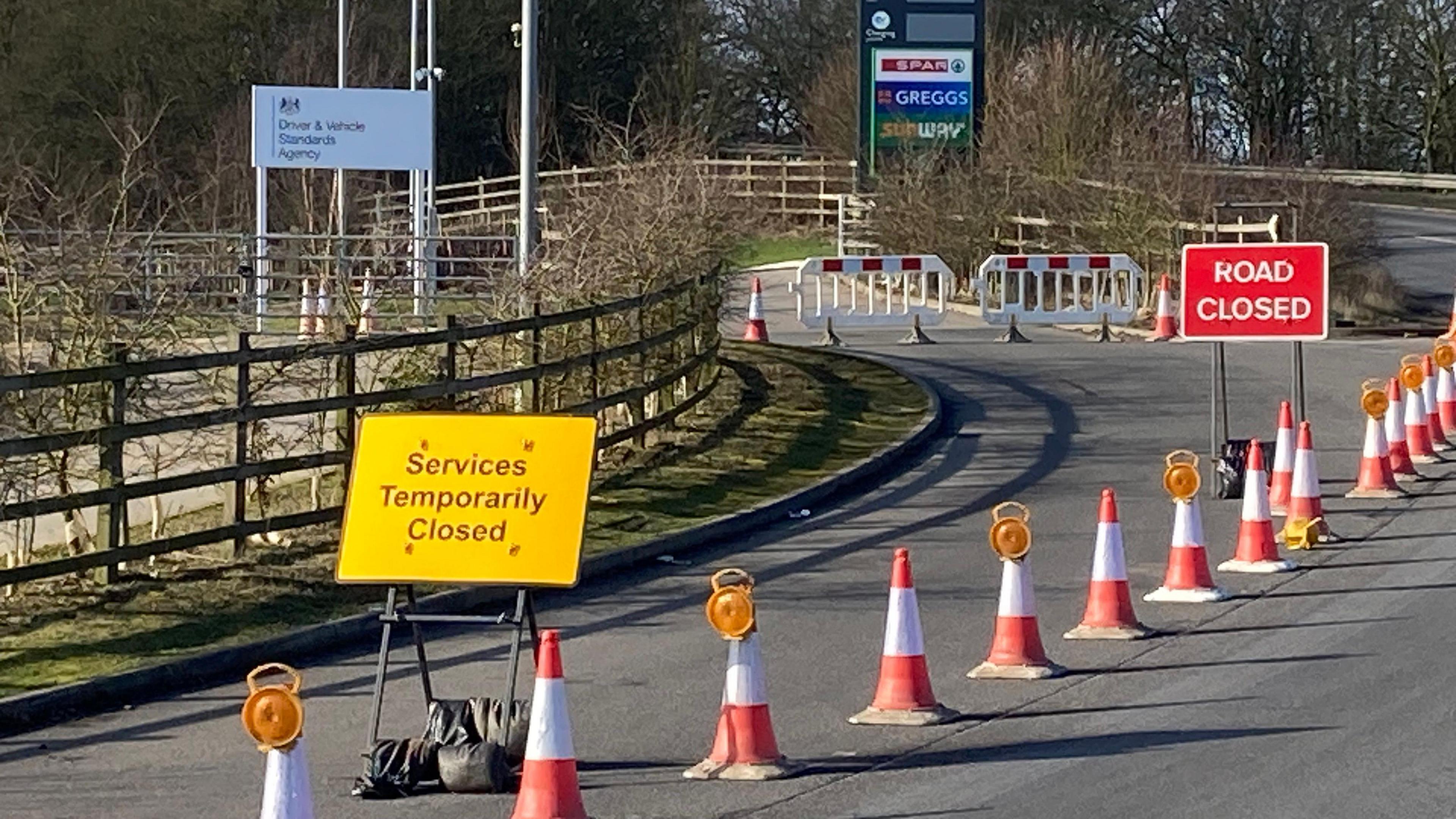 A line of cones have been put on the side of a road. There is a red sign that reads 'road closed' and a yellow sign that reads 'services temporarily closed.
The road is heading towards a service station