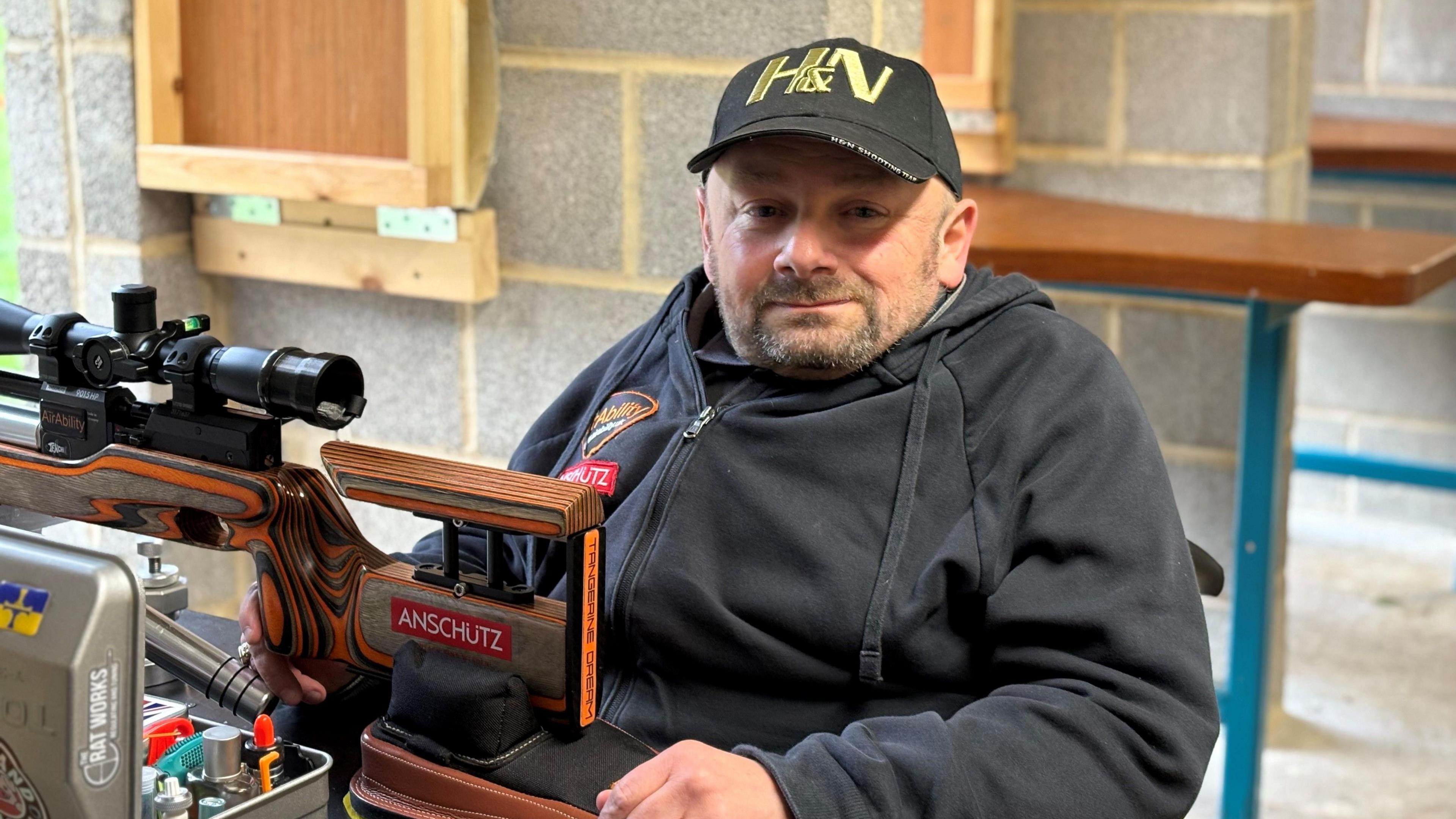 Matthew Gleaves wearing a cap and a black hoodie. He is sitting by an air rifle on the bench in front of him. 