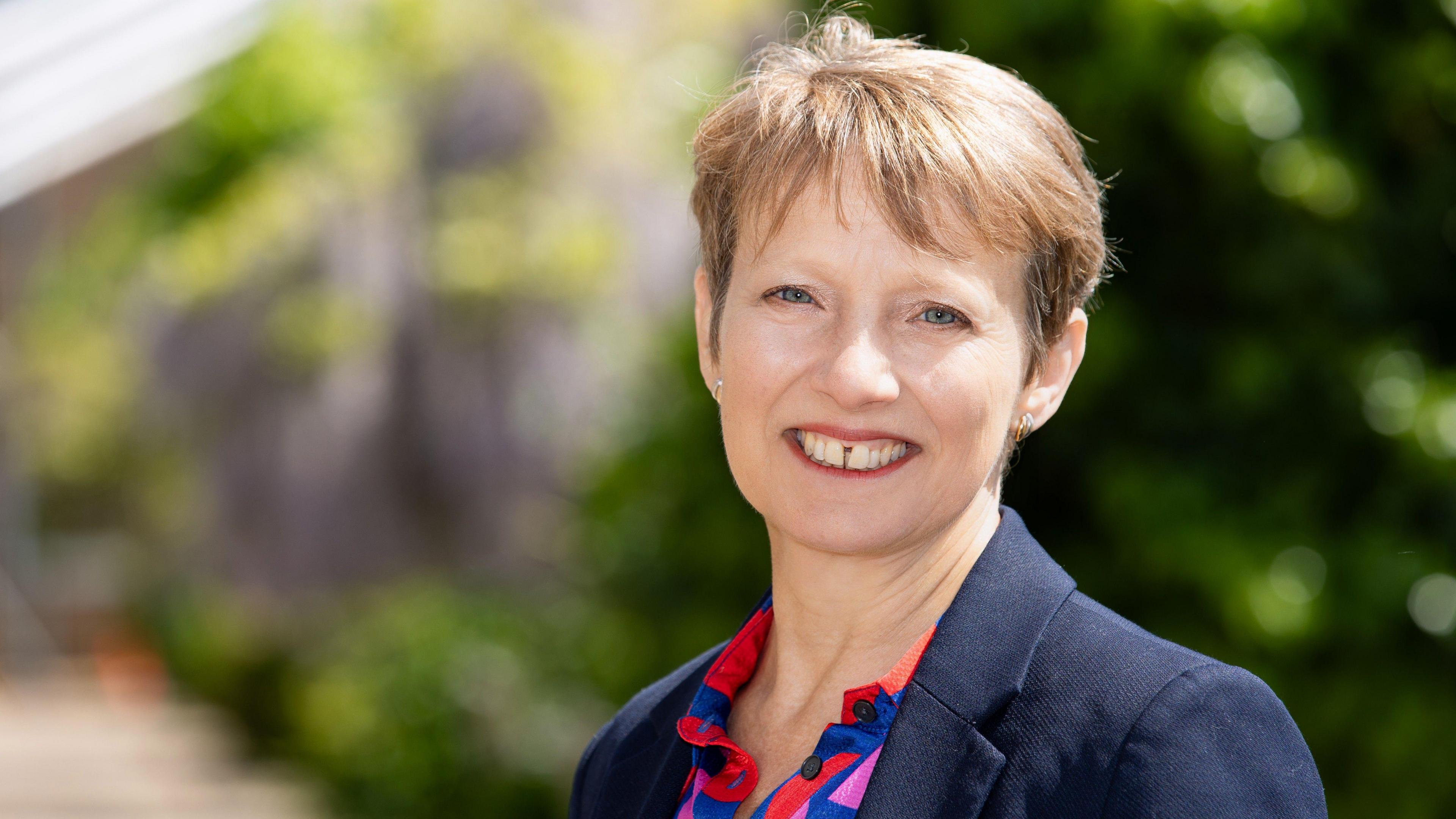 Clare Moody smiles at the camera in a navy blazer and collared patterned shirt. The background is blurred but it appears to be green foliage.